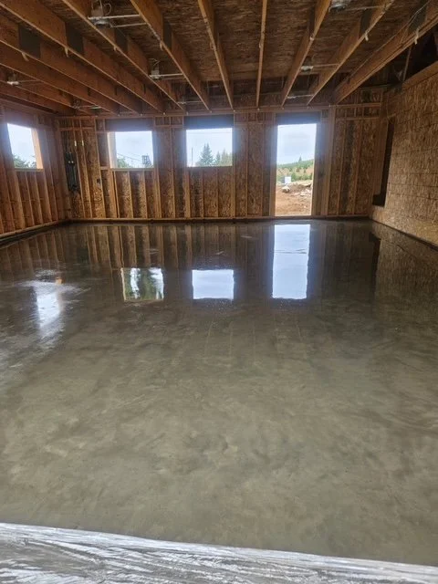 Construction site of a wooden building with a freshly poured concrete floor, exposed ceiling beams, and windows revealing outdoor scenery.