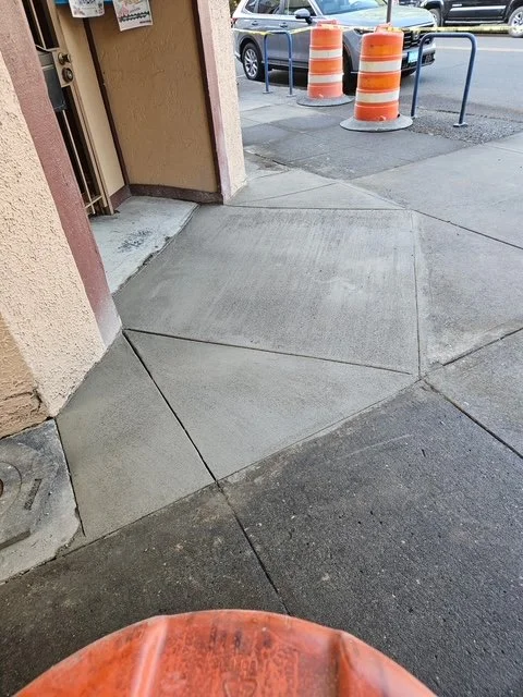 Sidewalk with two orange traffic barrels in a parking lot, a black car, and a building entrance.