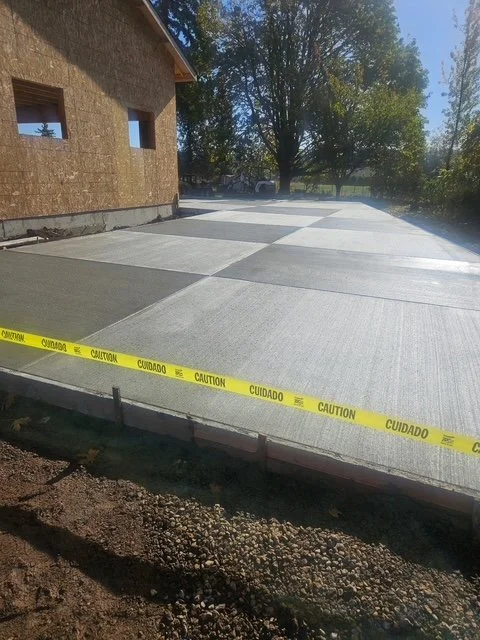 Newly poured concrete driveway with caution tape in front of a house under construction, surrounded by trees and dirt.