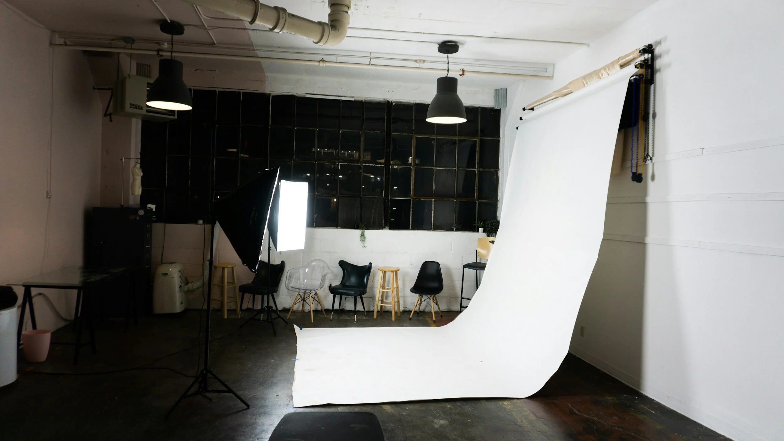 Photography studio with a white seamless backdrop, studio lighting equipment, chairs, and a large grid window in a loft-style space.