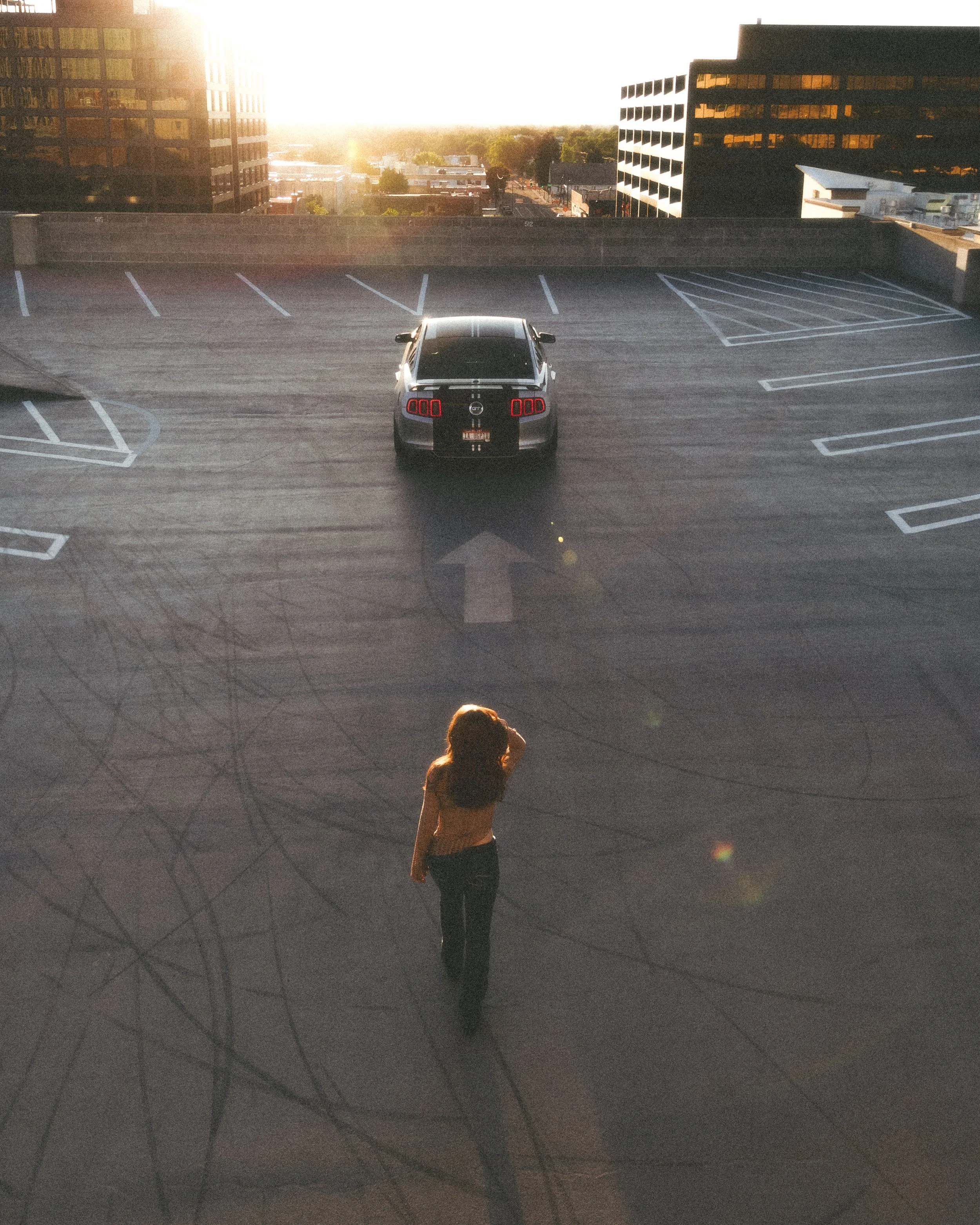 A young woman in dark clothing stands on the ground of an empty parking garage, with her right hand raised to her face, looking into the distance. The sun is setting behind tall buildings, casting a bright glow and long shadows across the scene.