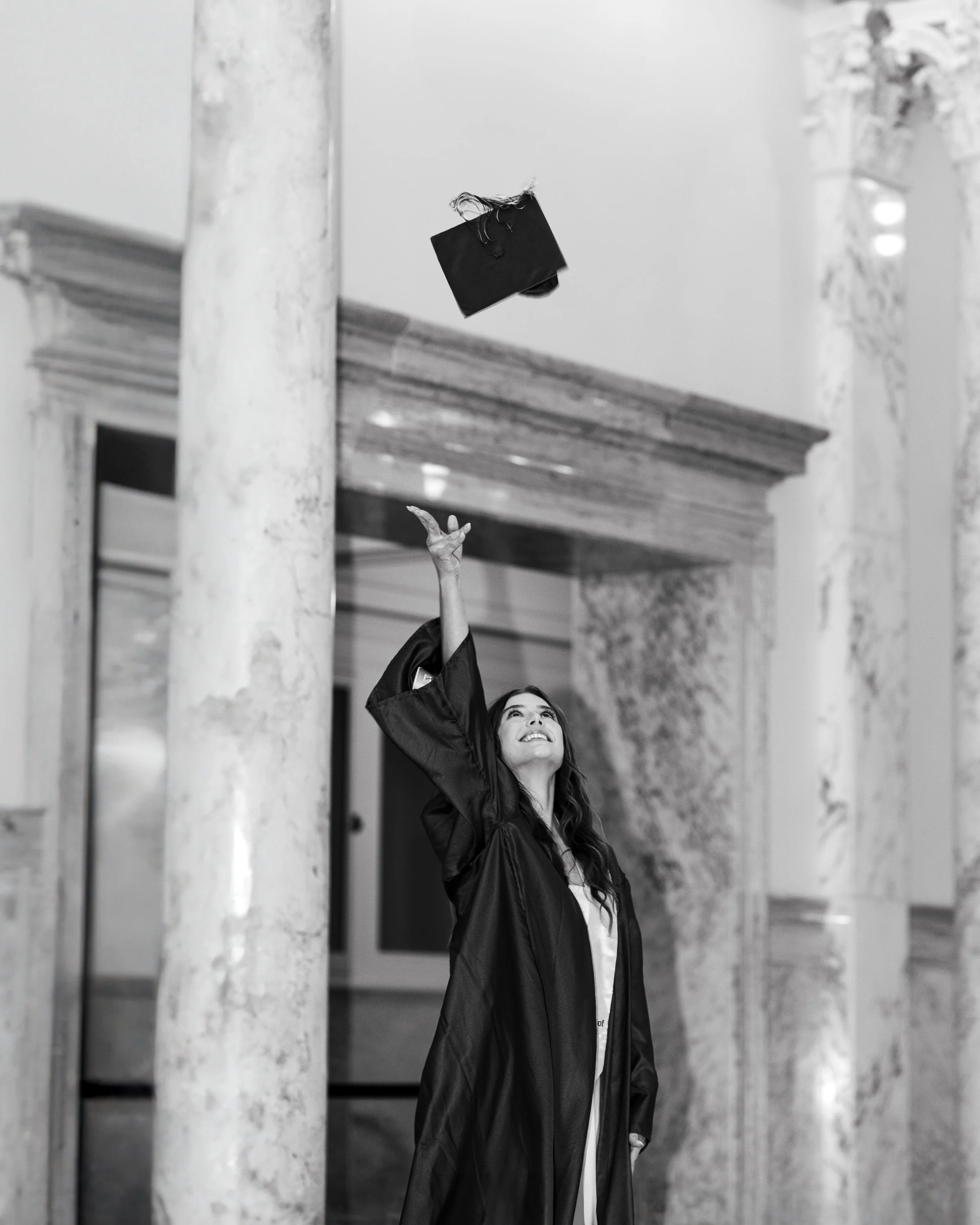 A young woman with long dark hair in her senior cap & gown is seen smiling and reaching up with her right hand as she catches her graduation cap as it comes back down. Taken in the Boise Capital Building.