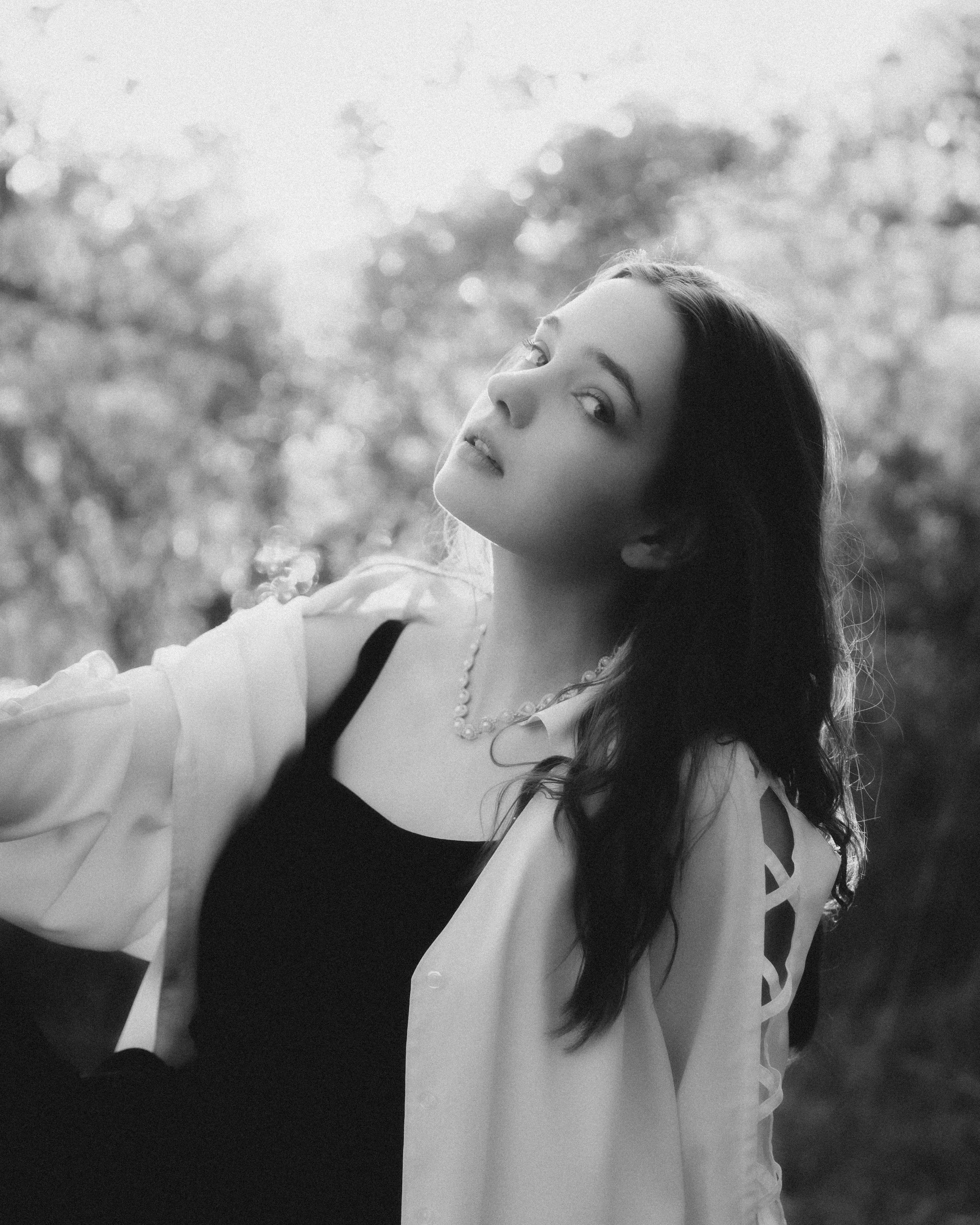 Black and white photo of a woman with long hair, wearing a white shirt with rolled-up sleeves and a beaded necklace, looking over her shoulder outdoors with trees blurred in the background.