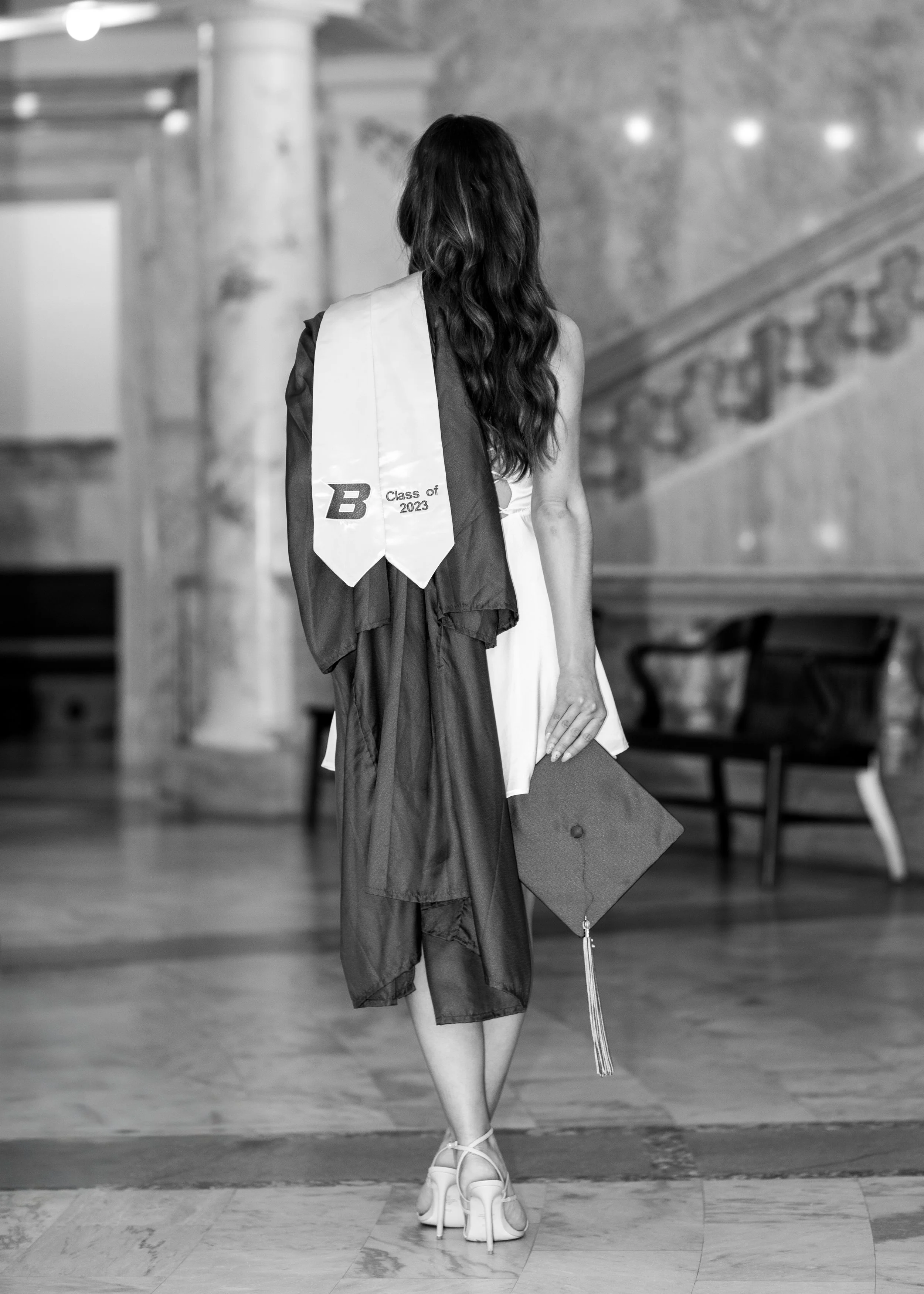 A young woman in a graduation gown and cap, holding her cap beside her, standing indoors with a staircase and bench in the background.