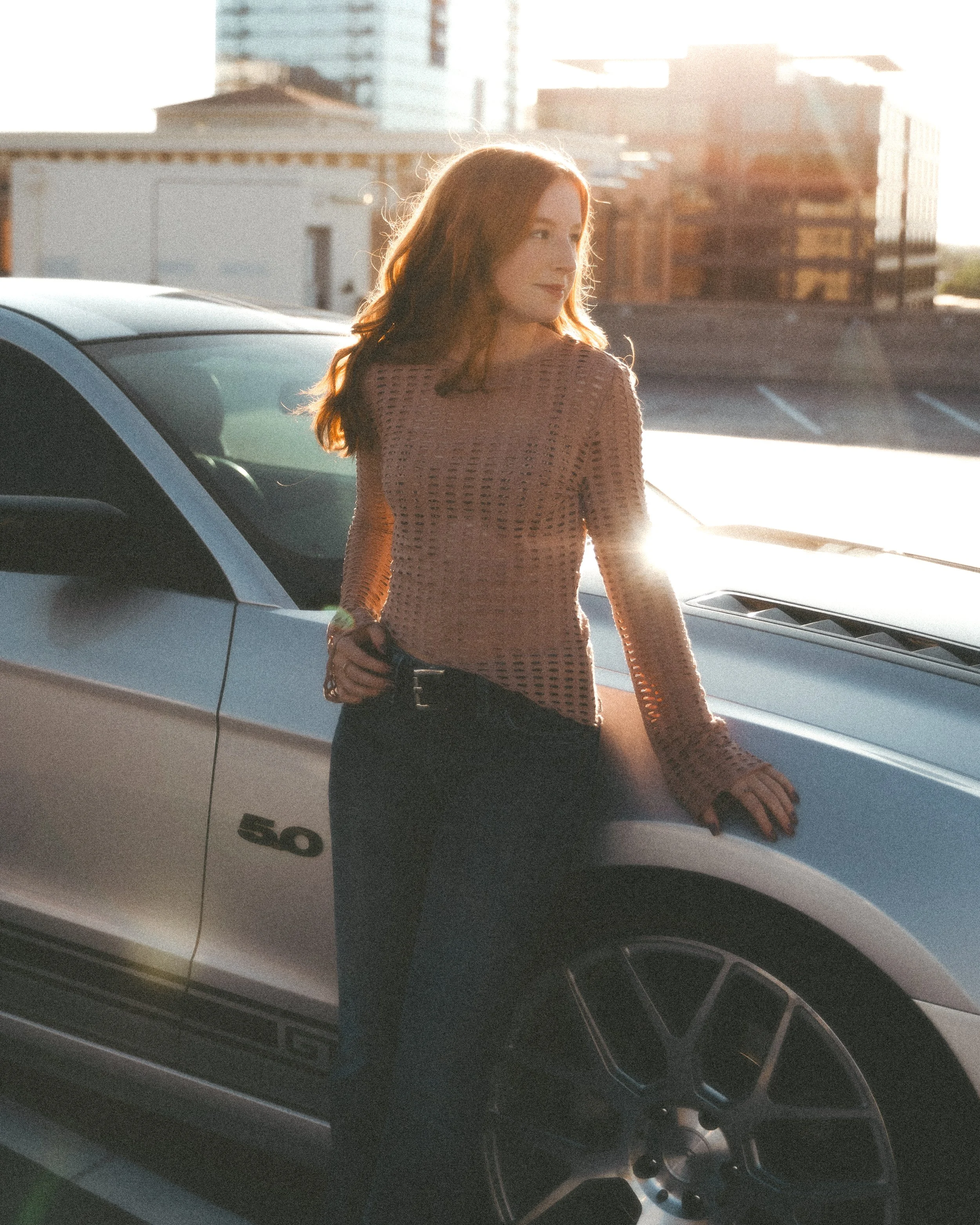 A woman with red hair standing in front of a white car, with a cityscape in the background during sunset.