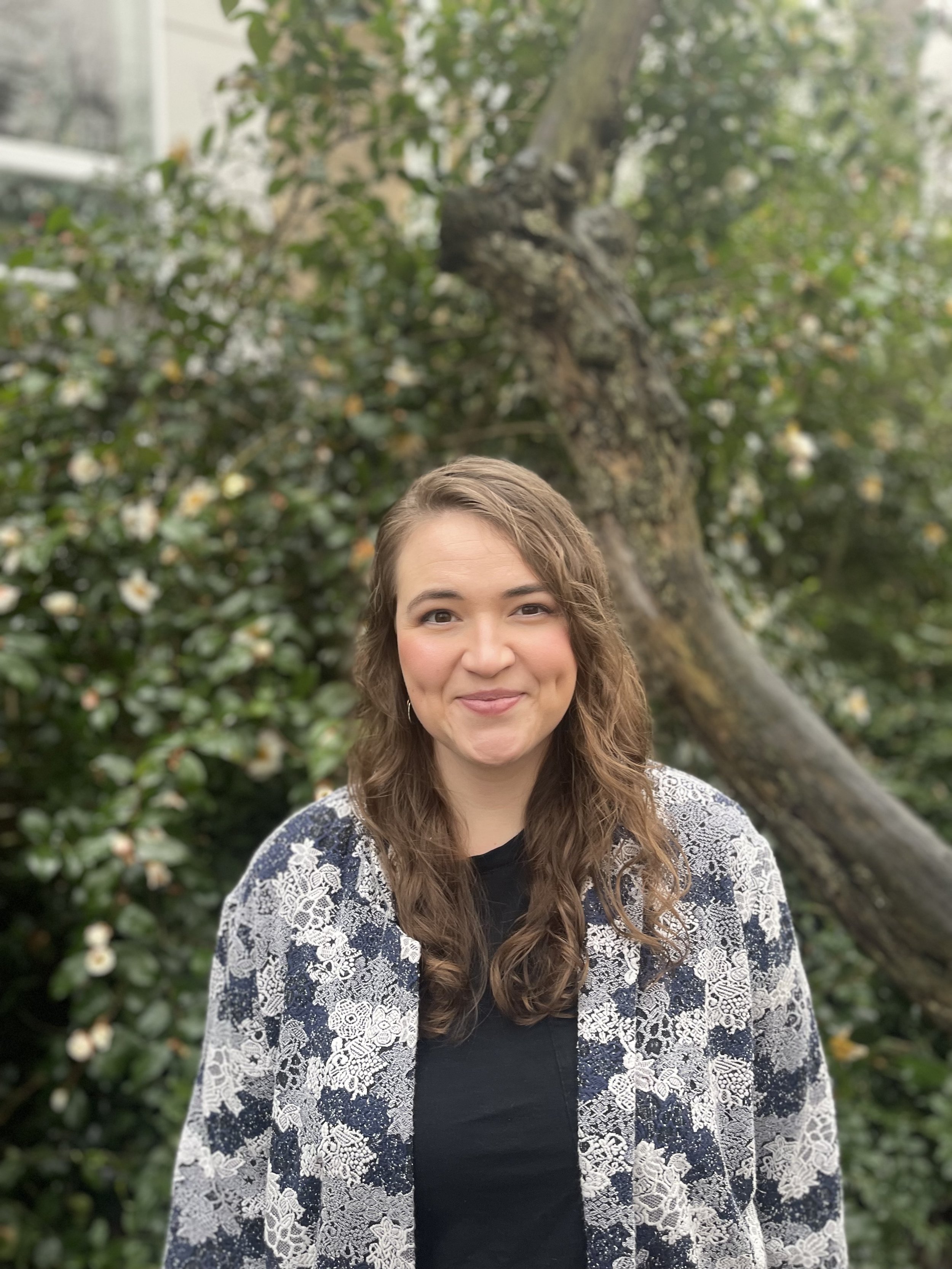 Young woman with long brown hair smiling outdoors in front of a tree with white flowers, wearing a black top and a lacy blue and white cardigan.