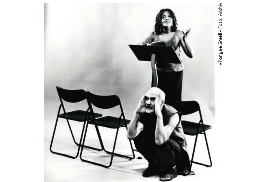 A woman standing at a music stand gesturing while a man is crouched in front, holding his head with his hands, with empty chairs around them.