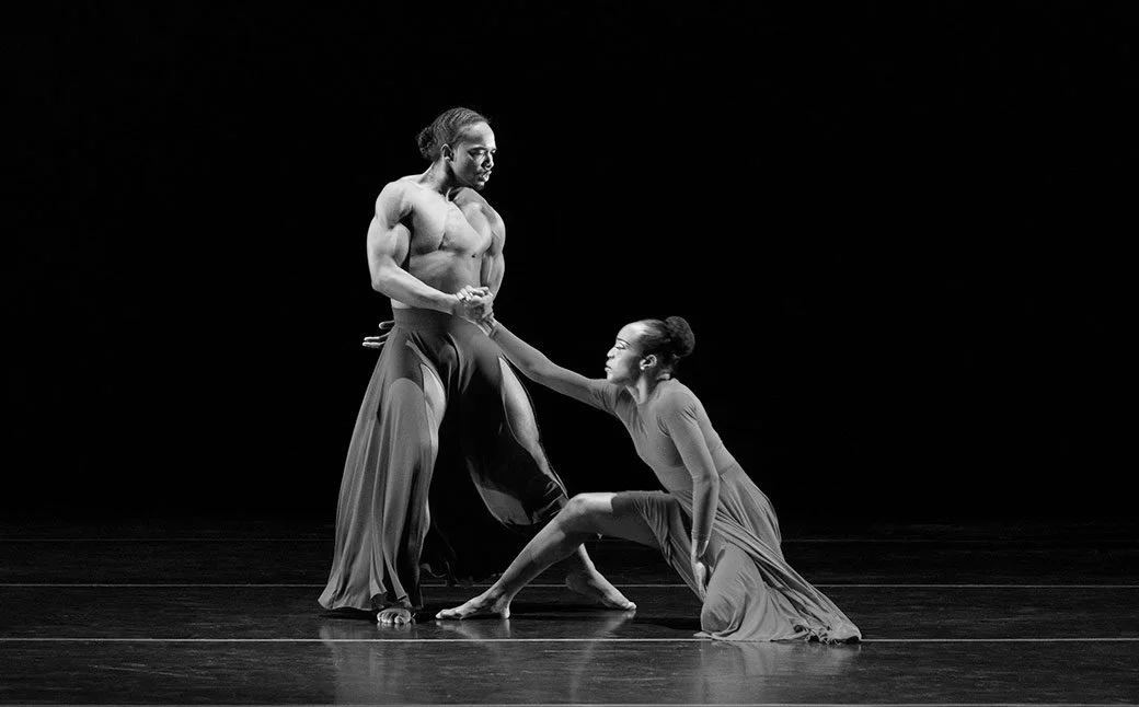 A black and white photo of a male and female ballet dancers on stage, with the male standing shirtless and wearing loose pants, and the female kneeling in a dress, engaging in a dance pose.