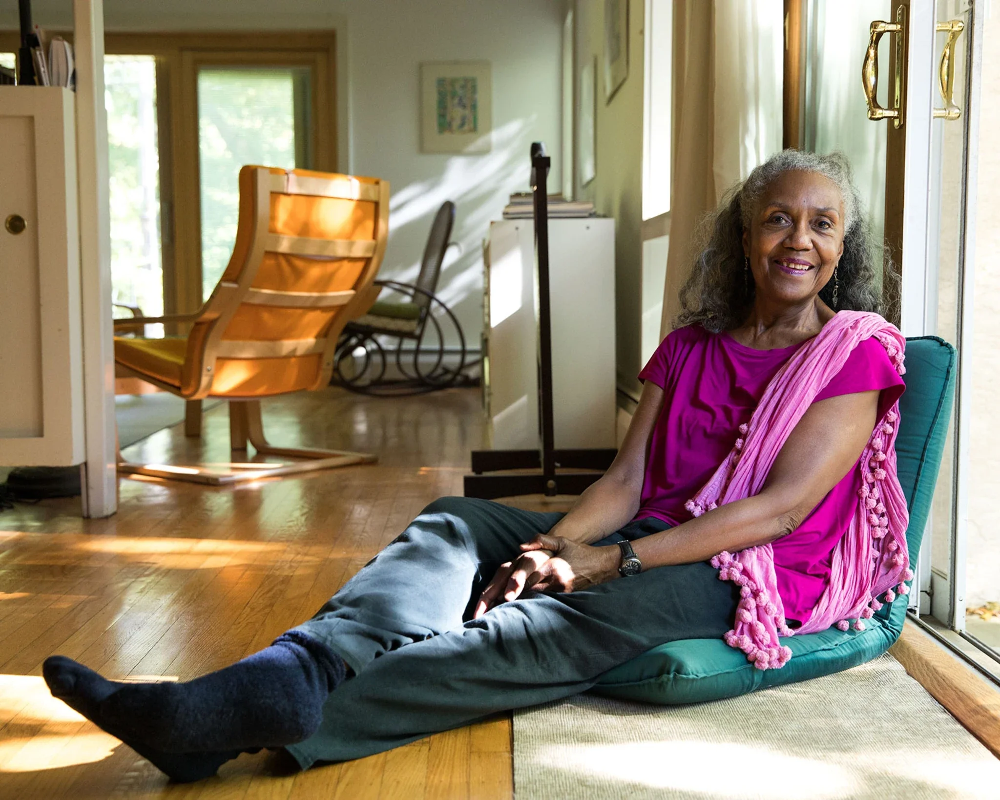 An elderly woman with gray hair, dressed in a pink top and wrap, sitting on a couch by a glass door, smiling at the camera. The room has wooden floors, and there are chairs, books, and artwork on the walls. Sunlight is streaming in through the window.