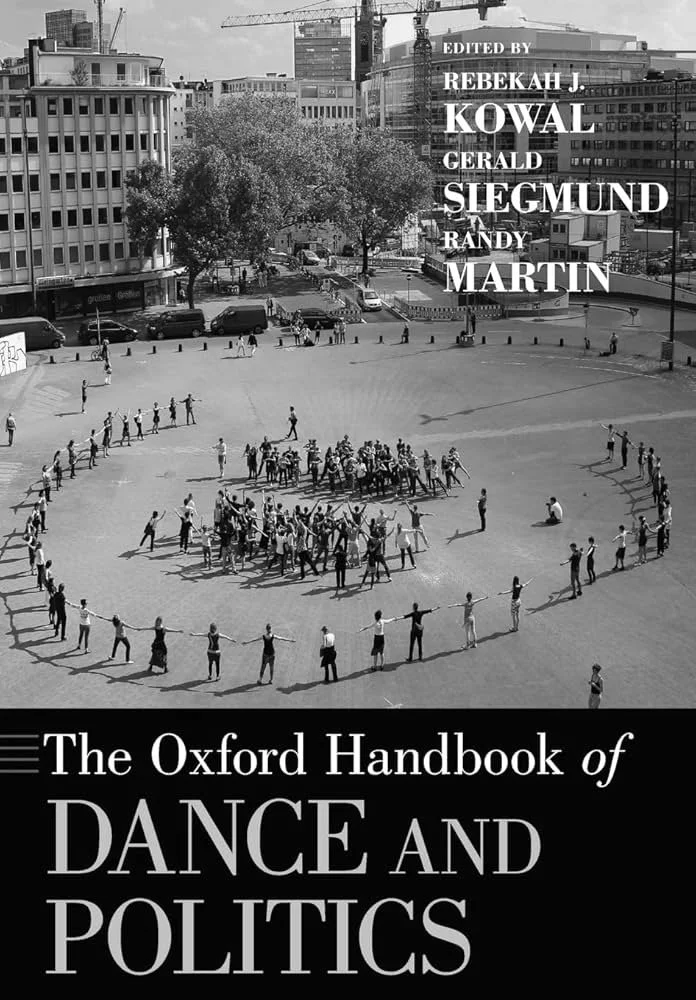 Black and white photograph of a large group of people forming a circle in an open urban area, surrounded by buildings and trees, with some people dancing or holding hands.