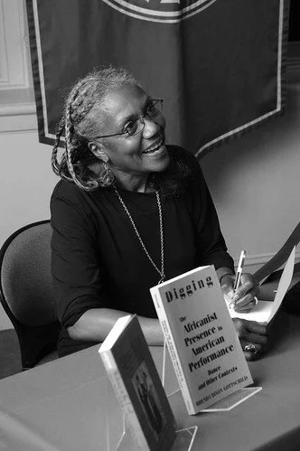 A woman with glasses and long gray hair smiling at a book signing event, sitting at a table with books and signing a copy.