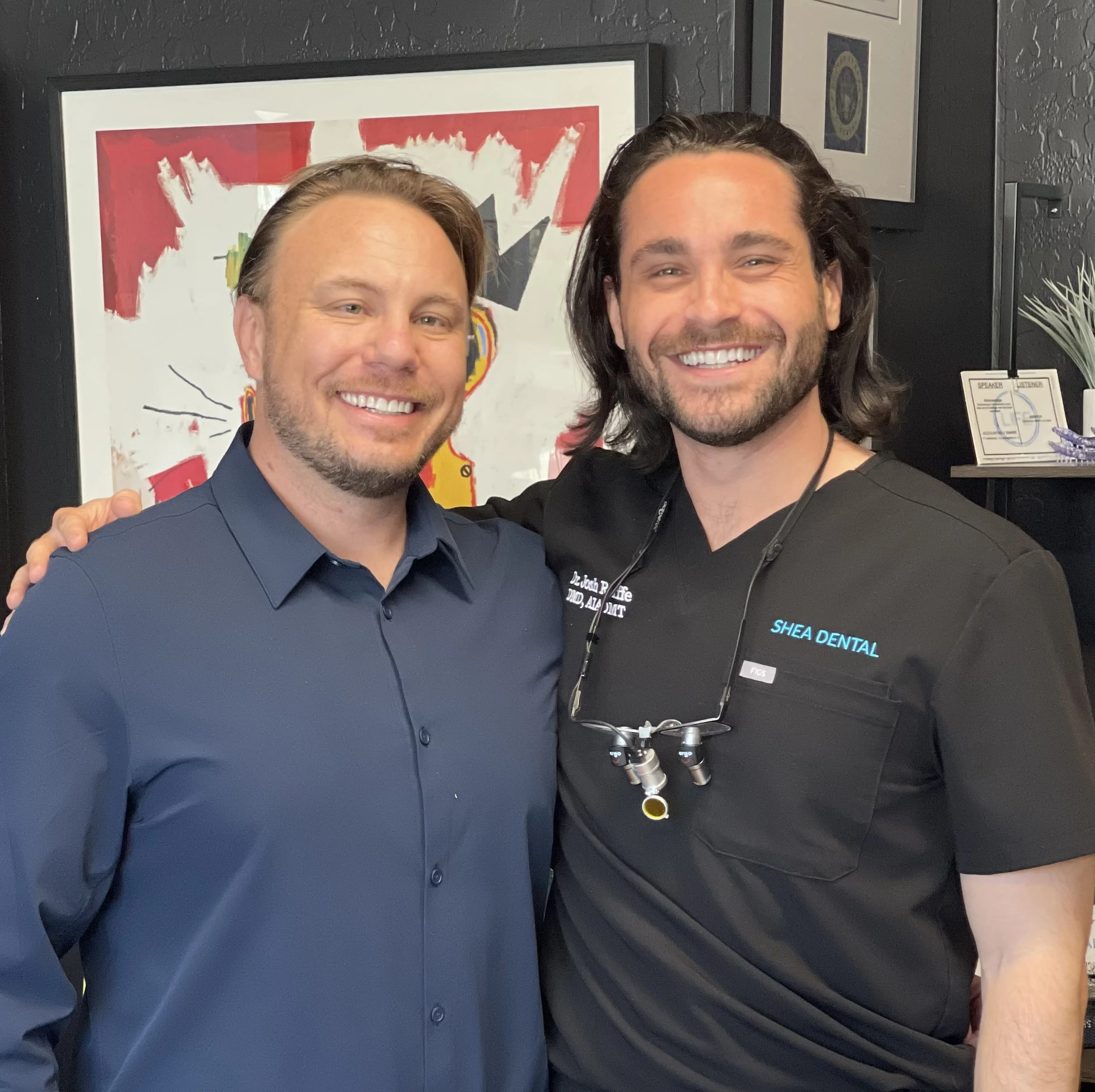 Two men smiling for a photo at an indoor event, partially visible framed artwork on the wall, one wearing a dark blue collared shirt, the other in black scrubs with a name tag that says Shea Dental.