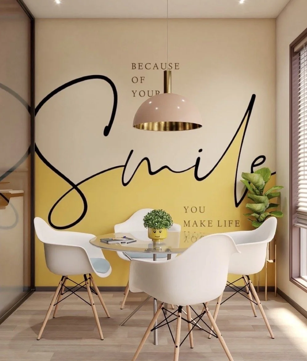 Decorative orthodontic dining area with a round table, four white chairs with wooden legs, a small yellow plant pot with a green plant, a large wall mural with the words "because of your smile you make life" and a large pink hanging light fixture.