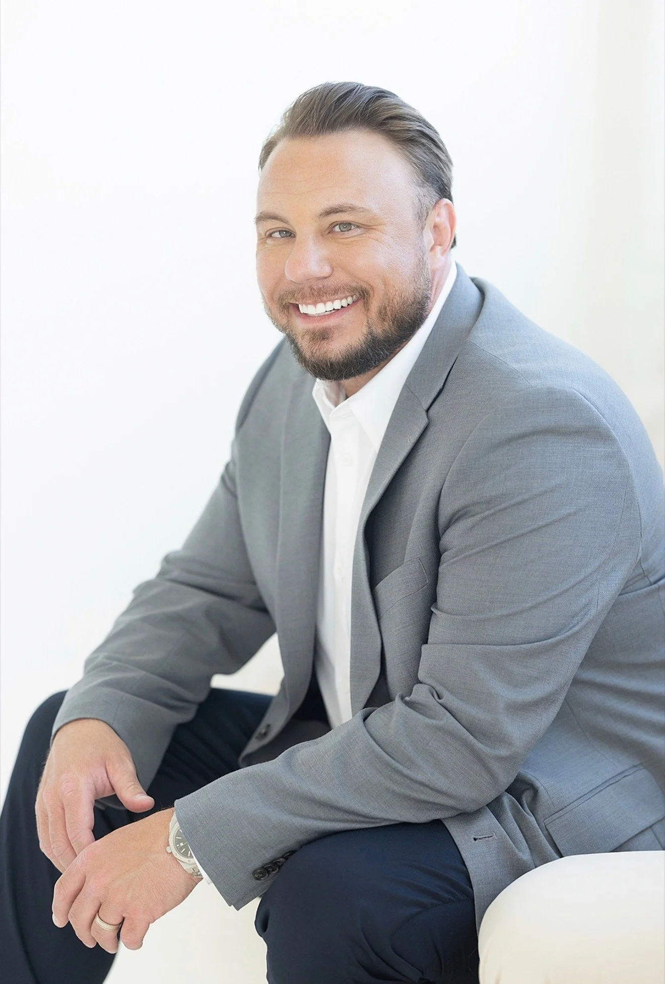 A smiling man with a beard and mustache wearing a grey suit jacket, white shirt, and a wristwatch, sitting and looking to the side against a plain white background.