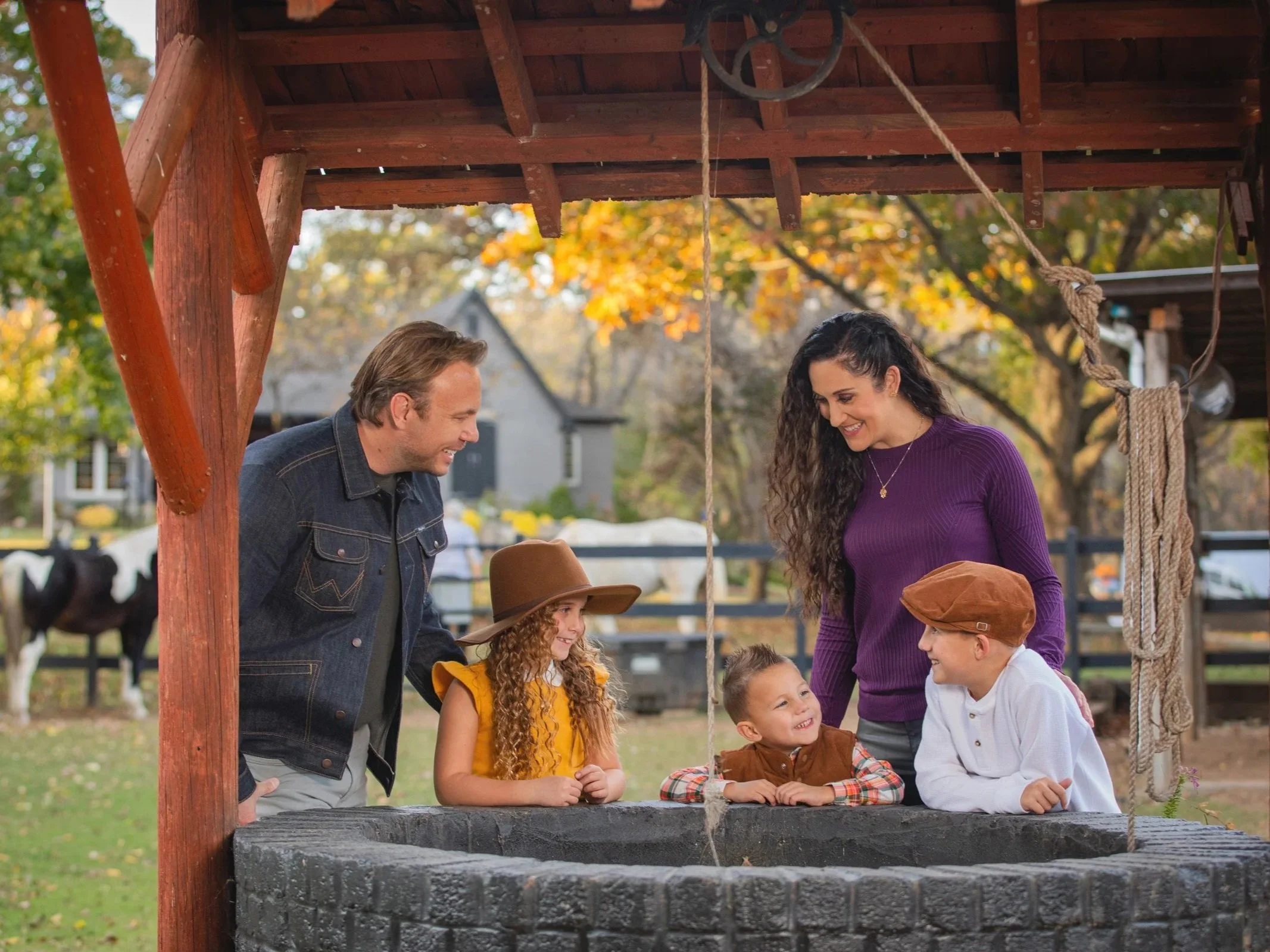 A group of children and adults gathered around a stone wishing well, engaging in conversation and smiling outdoors in a park with autumn foliage.