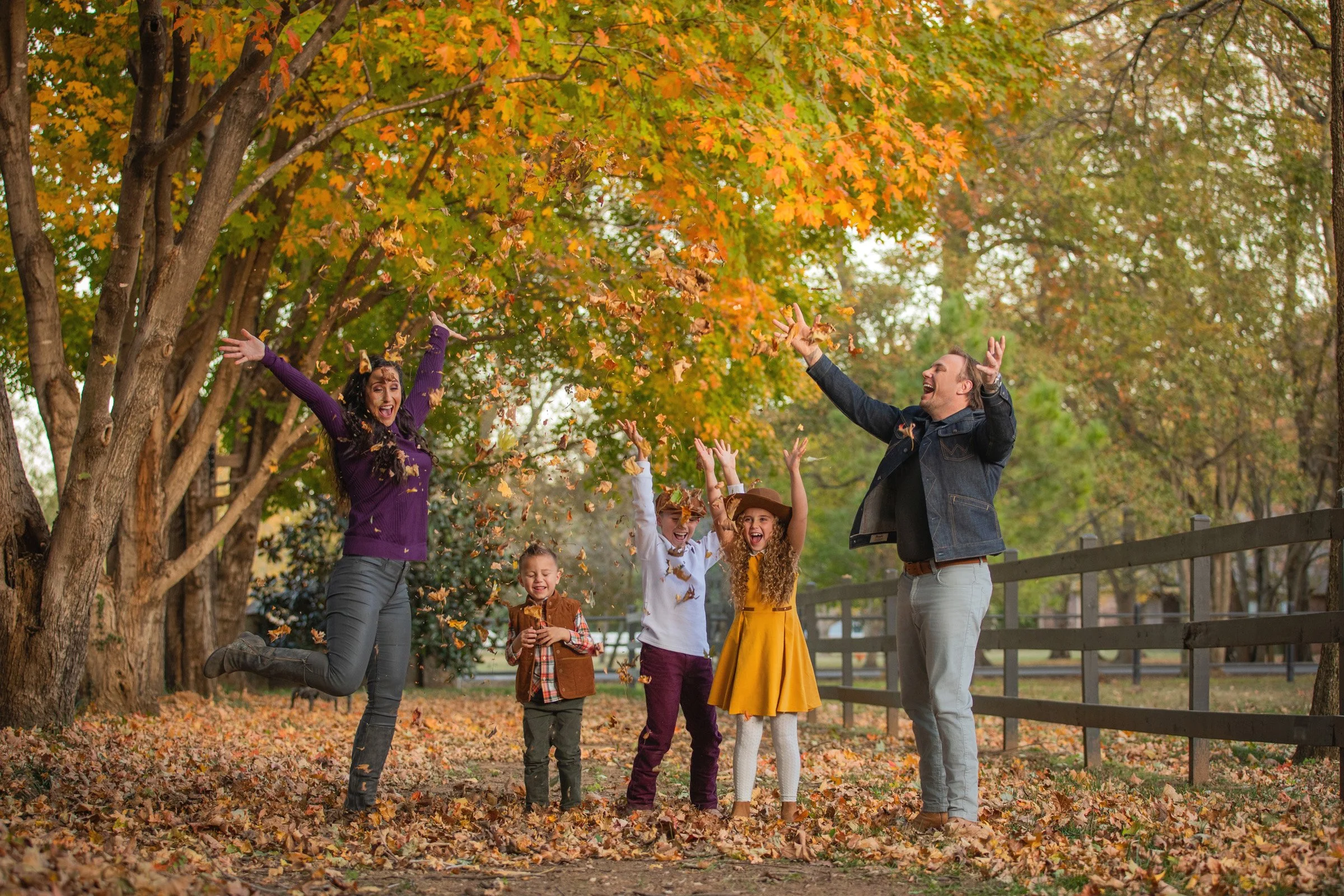 A group of children and two adults joyfully play among falling autumn leaves in a park, with trees displaying colorful fall foliage in the background.