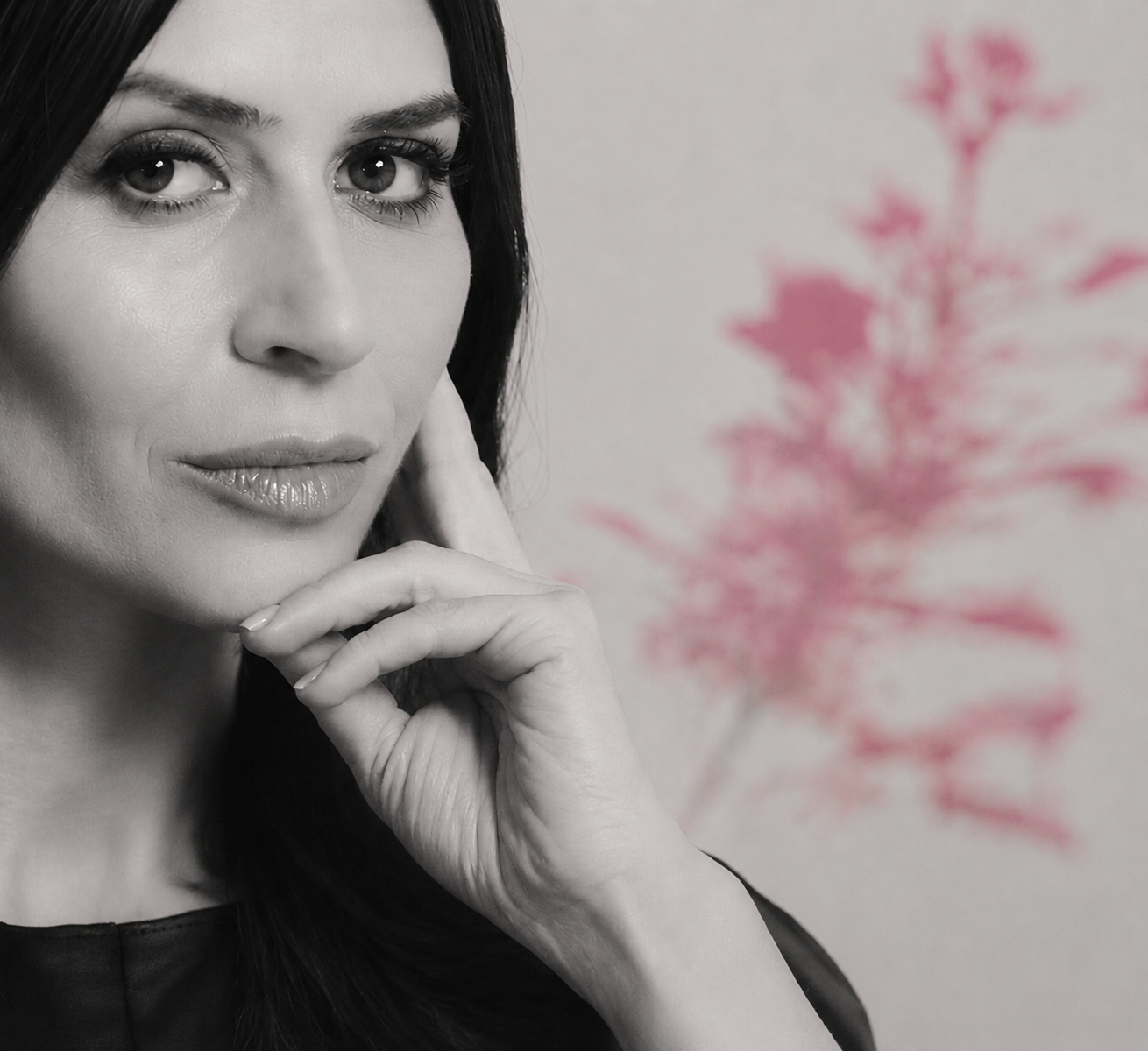 A woman with dark hair and light skin looking at the camera with her hand resting near her chin, with a blurred pink-red outline of a tree or plant in the background.