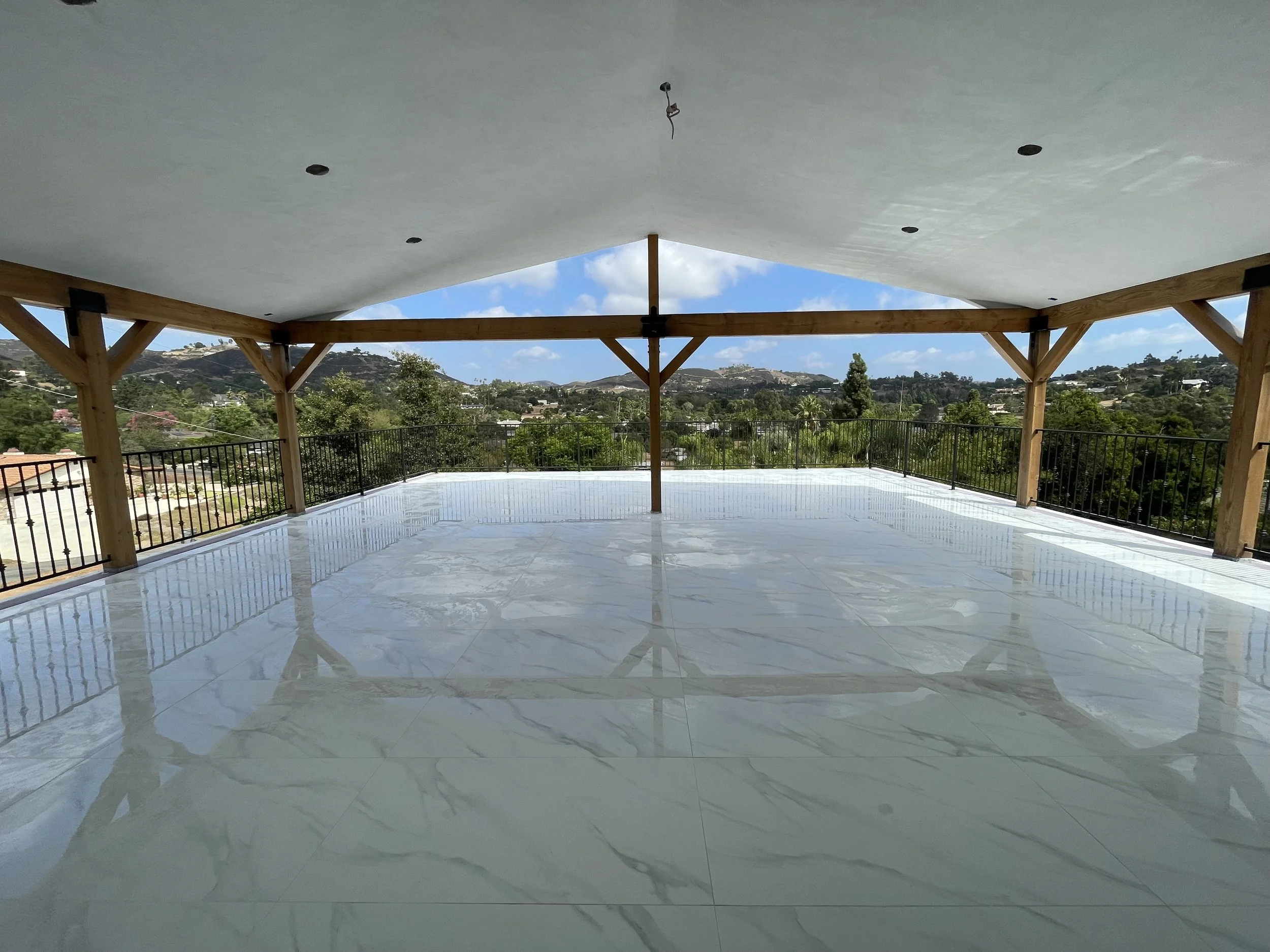 An outdoor space with a wooden beam structure supporting a large, flat, smooth white ceiling. The floor has shiny marble or tile with reflections. There is a metal railing around the perimeter, and a scenic view of hills, trees, and houses under a partly cloudy sky in the background.
