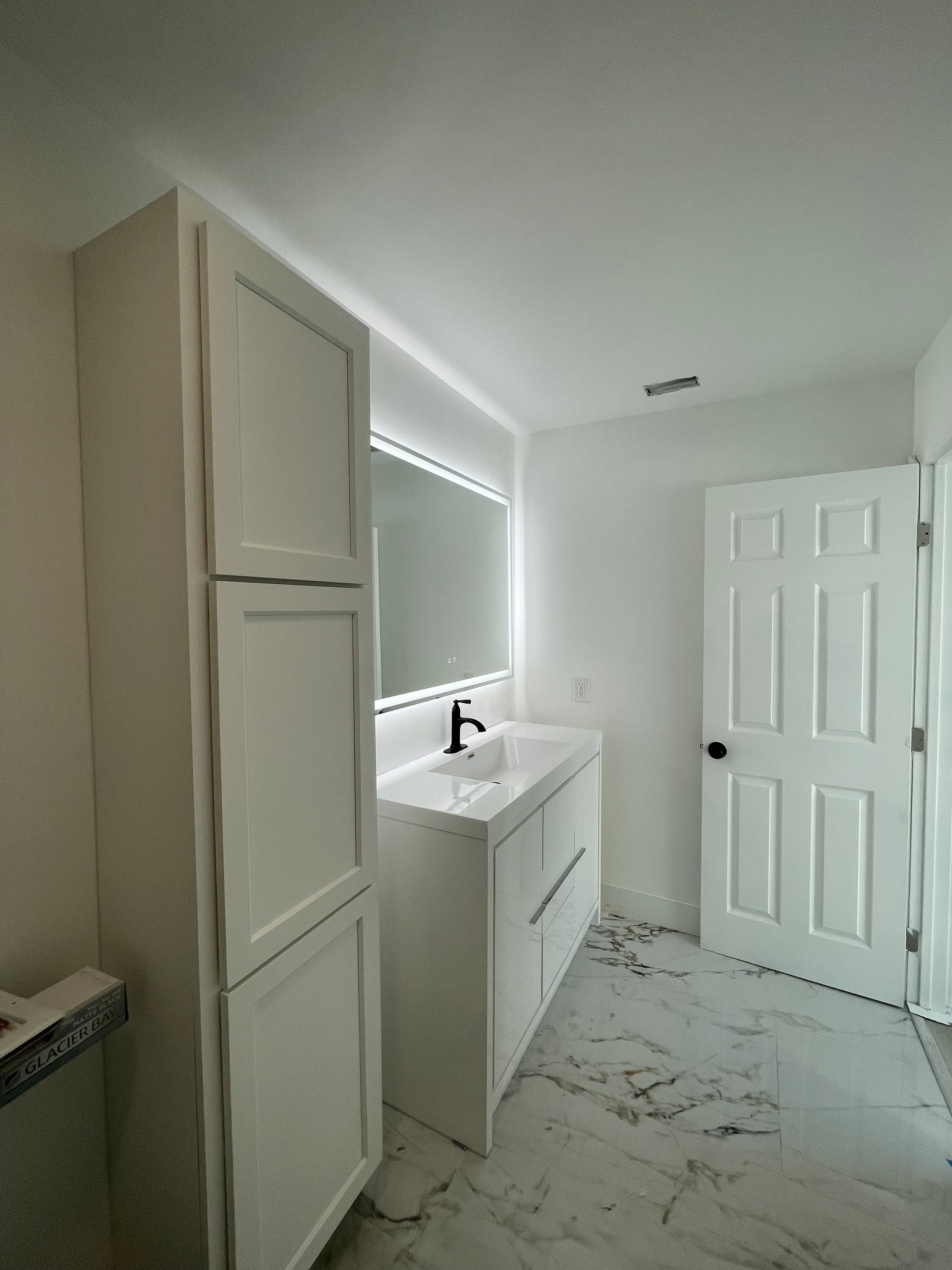 A modern bathroom with a white vanity, black faucet, white cabinets, mirror, white door, and marble tile flooring.