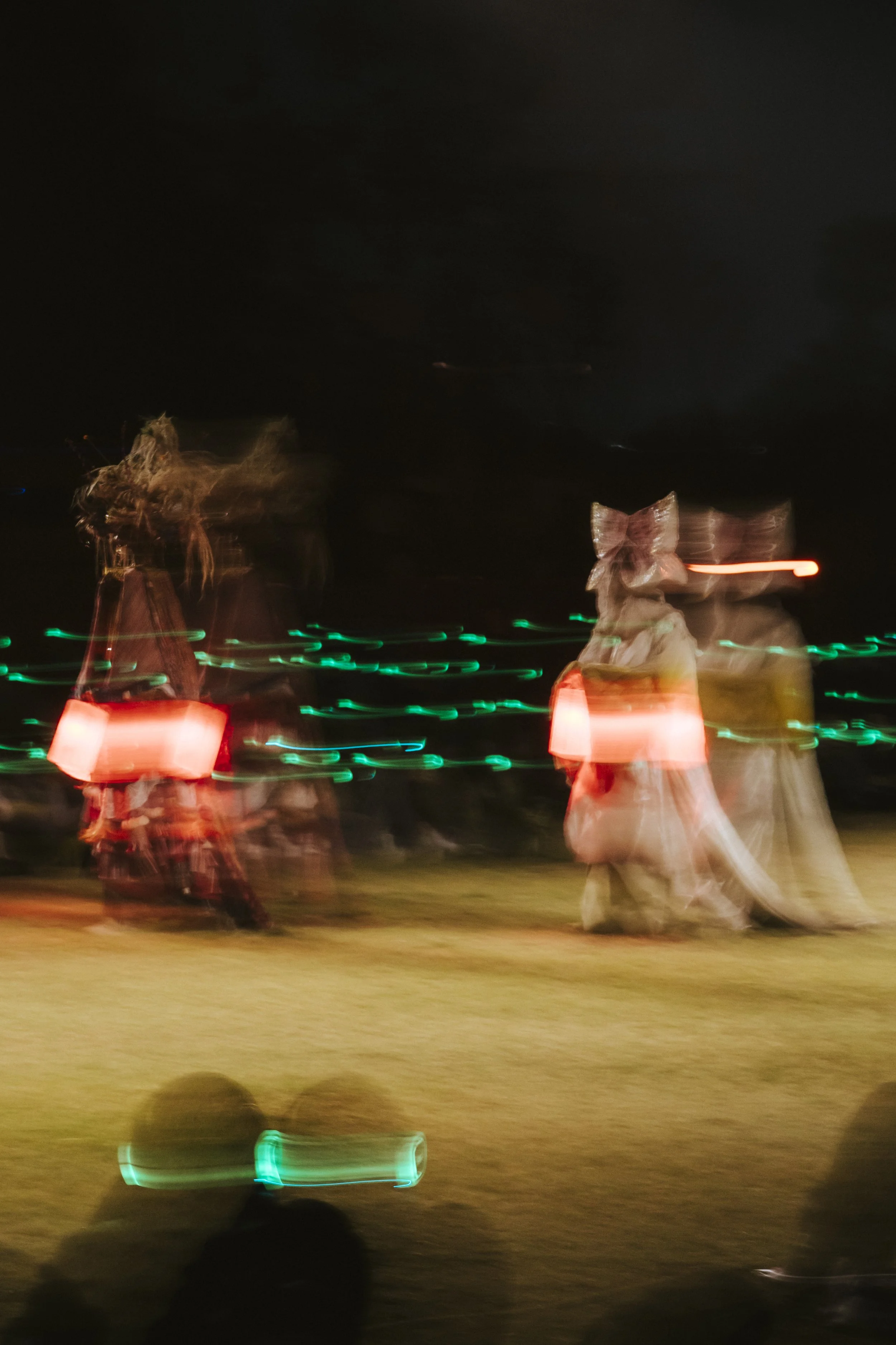 Two earthly costumed figures walking in the dark, carrying illuminated glowing lanterns, participating in a night parade. In the lower part of the image, there is an audience member wearing silent disco headphones looking on to the parade.