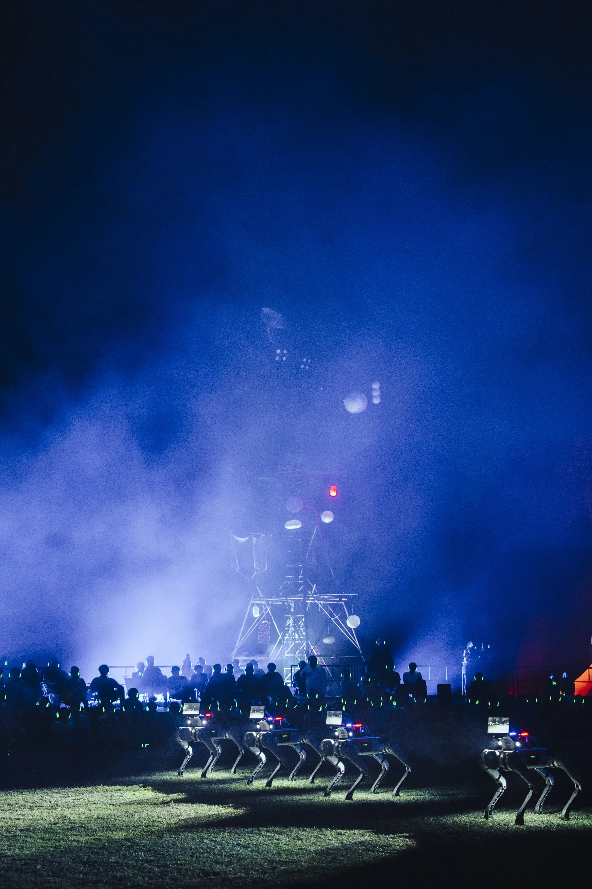 An outdoor stage with lighting and fog effects, with a group of audience members. In the lower part of the image, there are robotic quadruped figures standing menacingly in a line.