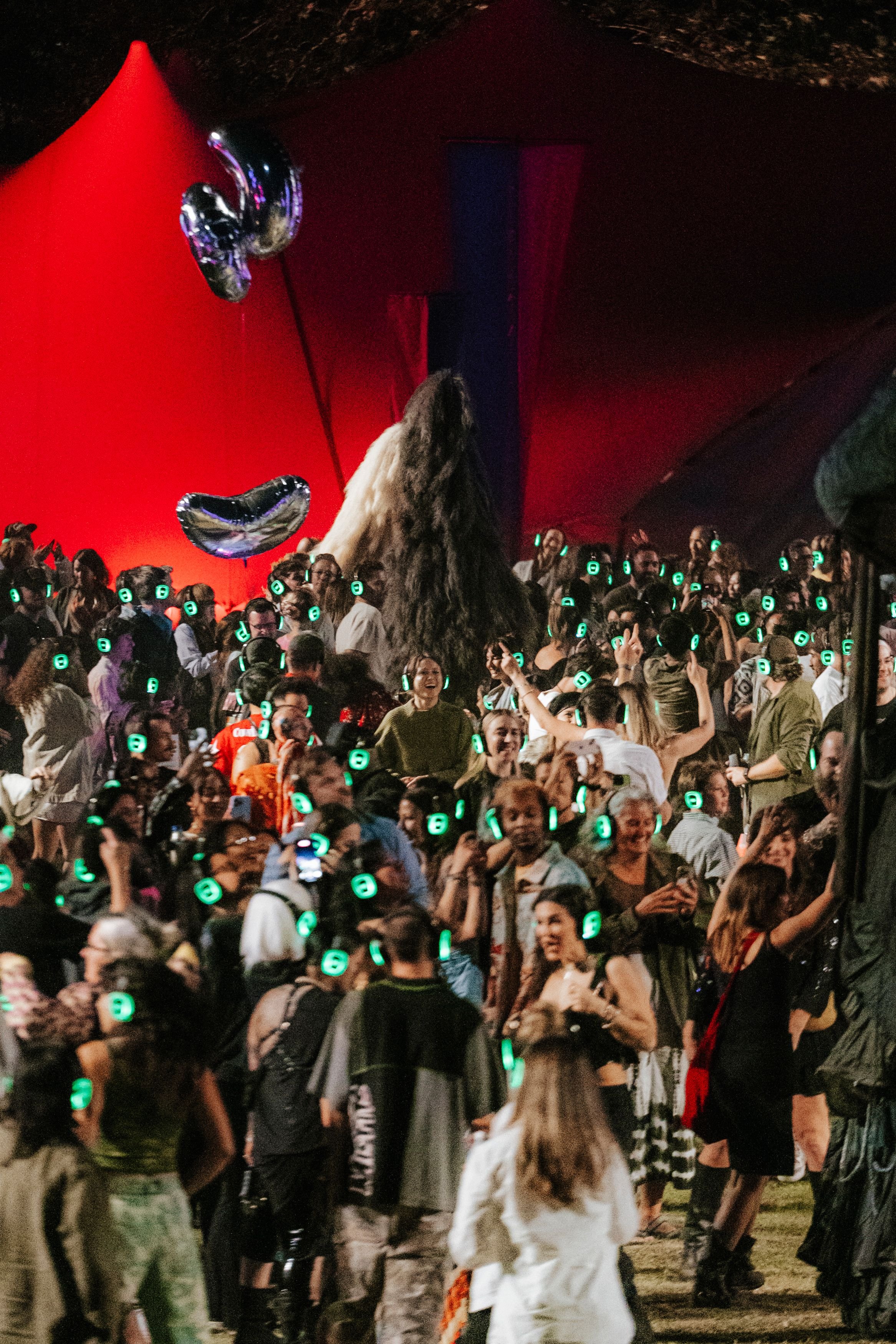 A large group of people wearing green silent disco headsets dancing and socializing outdoors before a large bedouin tent among puppets and robots adorned with silver mylar balloons.