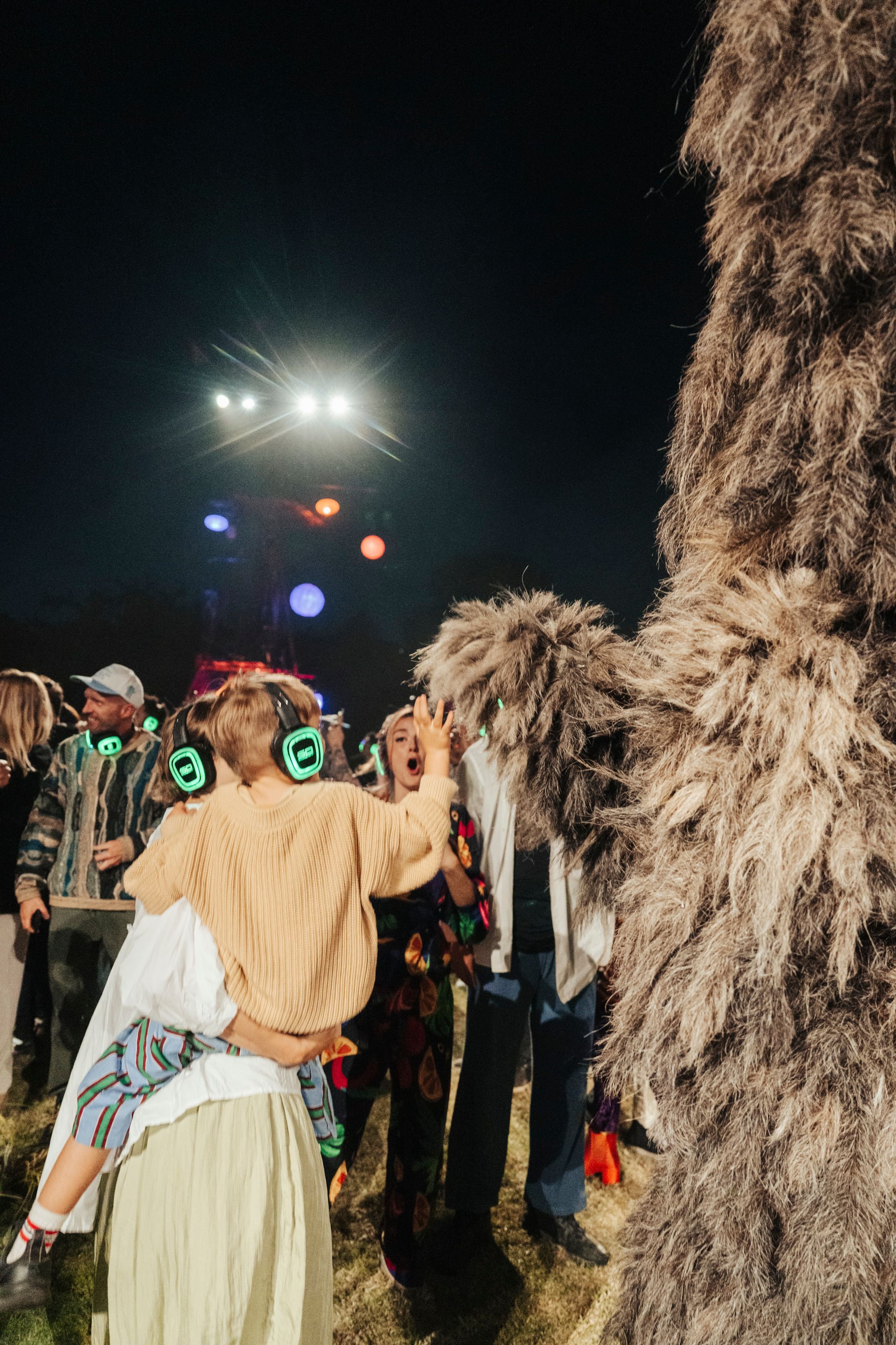 A group of people at an outdoor nighttime event, some wearing silent disco headphones. A mother and small child interact with a large archetypal puppet.