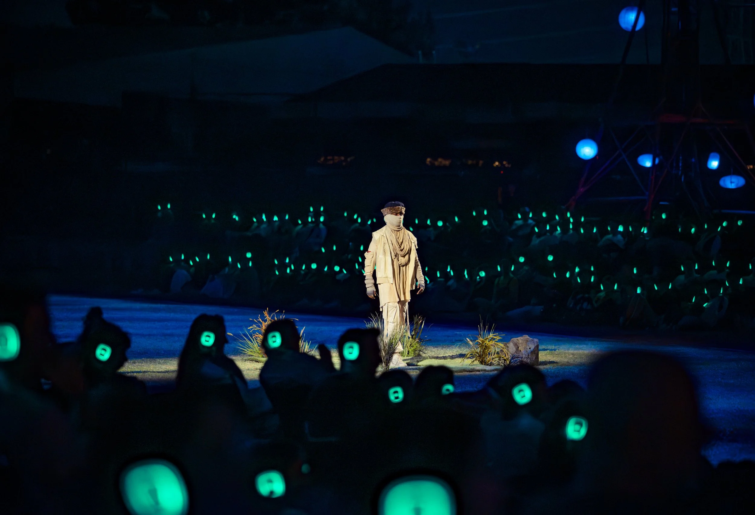 A an archetypal character dressed in ragtag global diasporic attire, including a head covering and face mask, walks alone on a dimly lit outdoor stage. Audience members in silent disco headphones look on and observe.