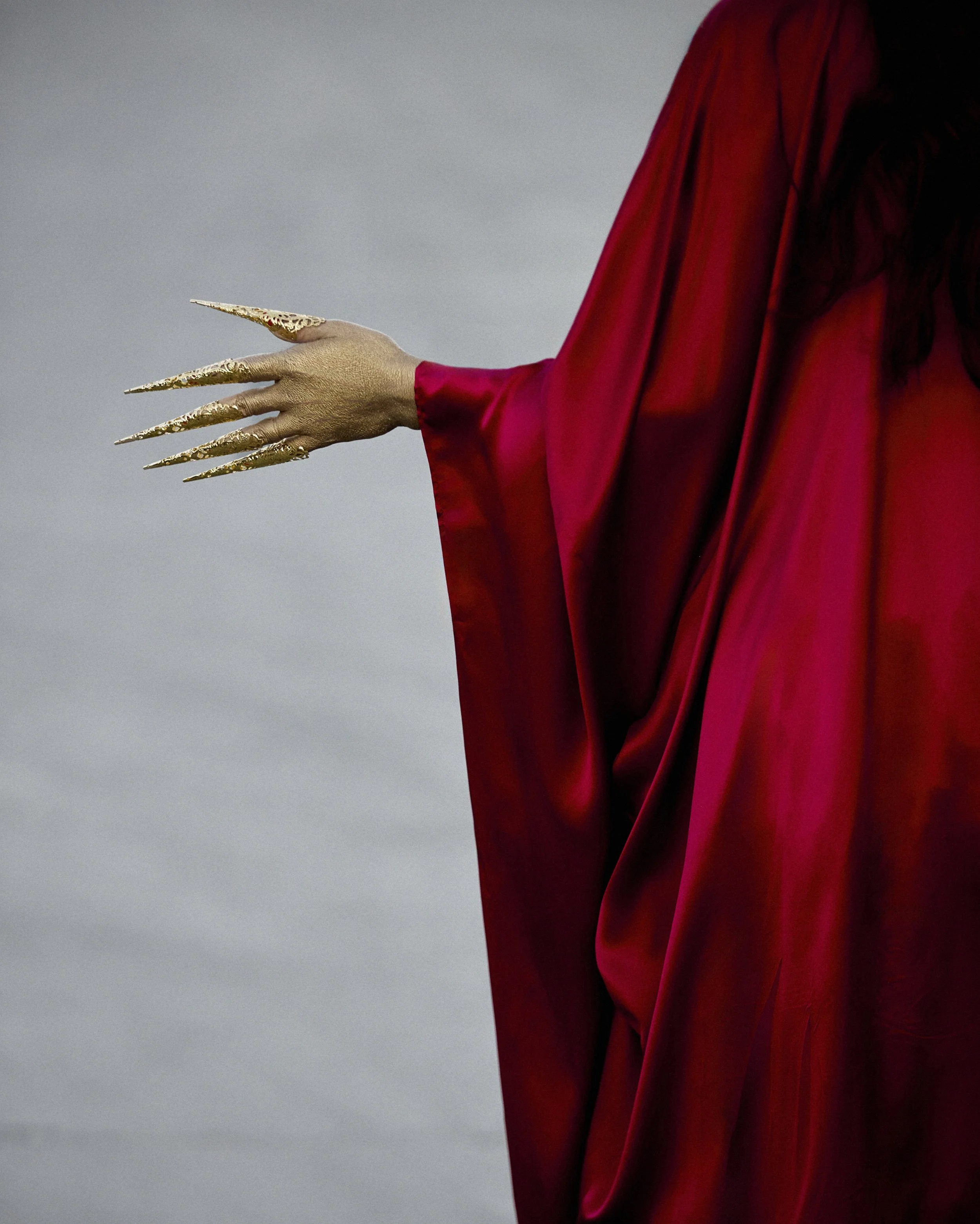 Figure wearing a red silk garment with a gold hand extended, fingers and nails elongated and laquered in gold.
