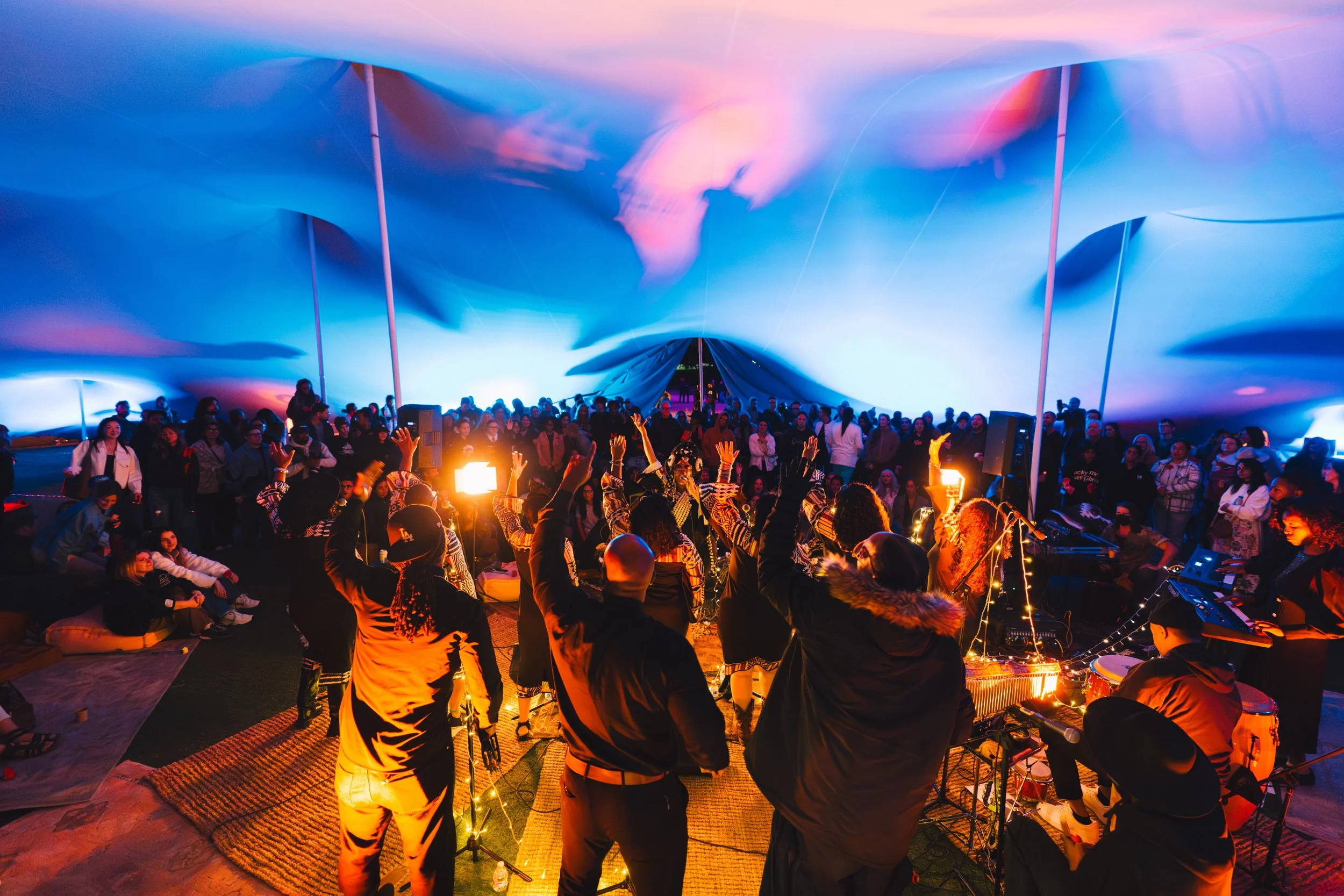 People dancing and enjoying live music inside a large, colorful, fabric-covered stretch event tent with vibrant pink and blue lighting.