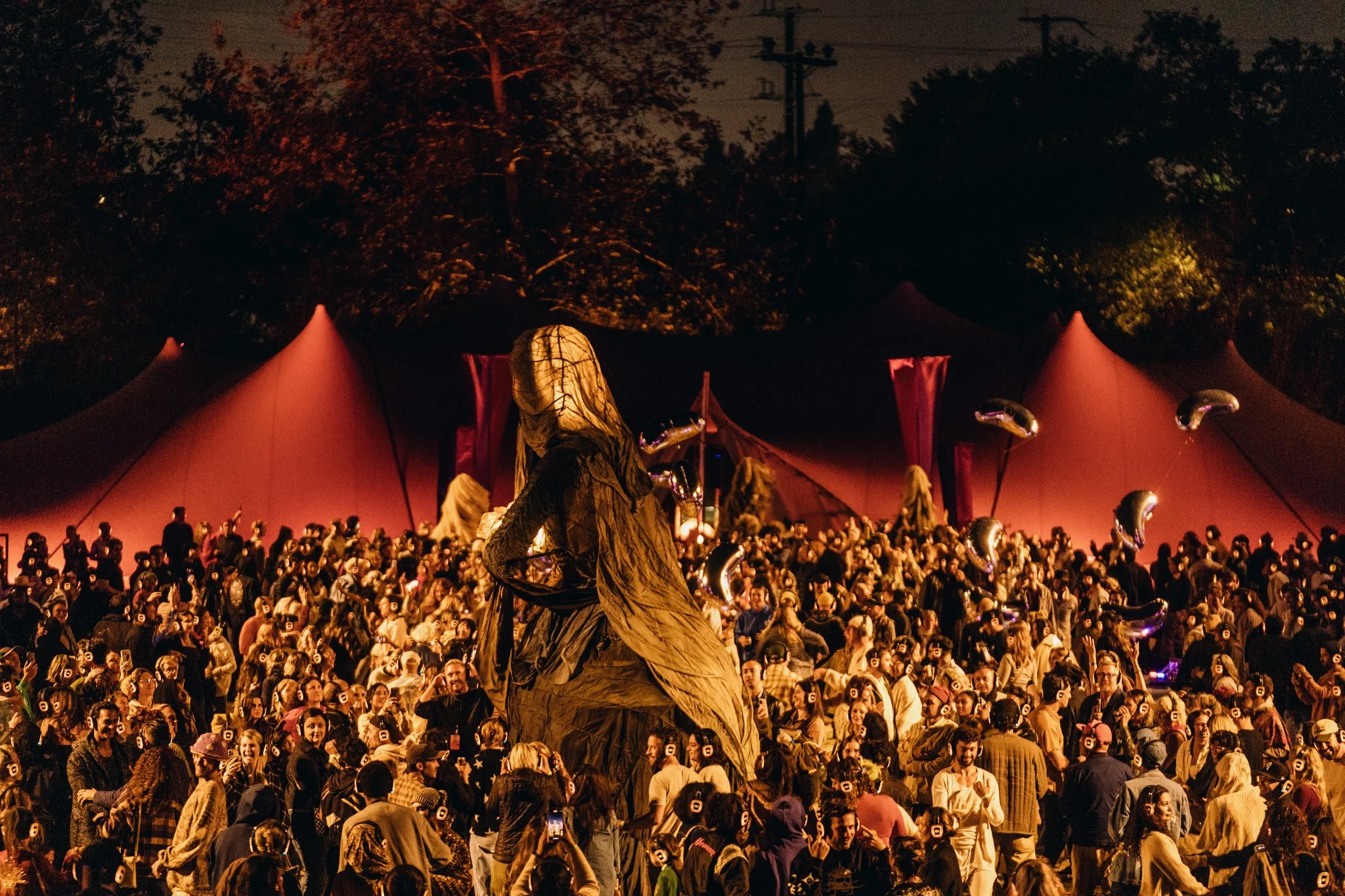 Large crowd gathering at night before a large tent. Participants are wearing silent disco headphones, dancing and socializing among large scale puppets and archetypal figures at The Cortège.
