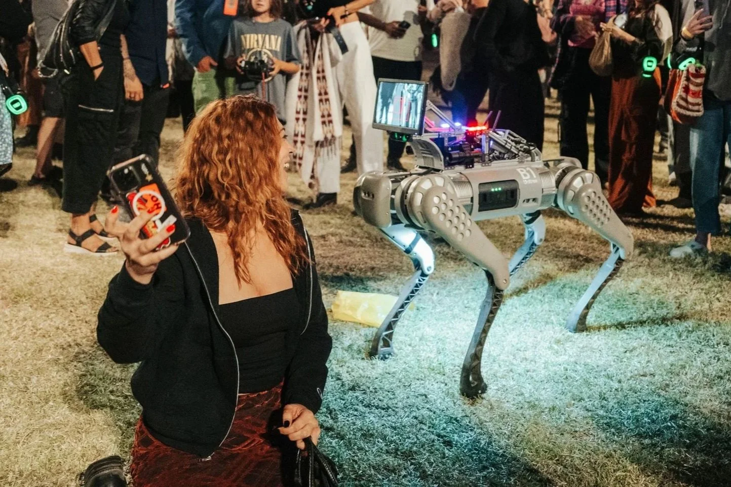 A woman kneeling on the grass, taking a selfie with a robotic quadruped with electronic components on its back.