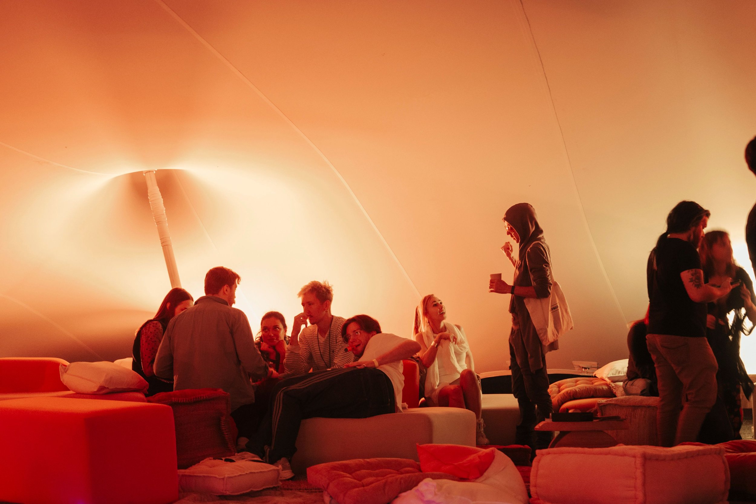 A group of people socializing and talking in a warm, orange-lit environment with large cushions and a low table, situated beneath the curved membrane of the stretch tent.