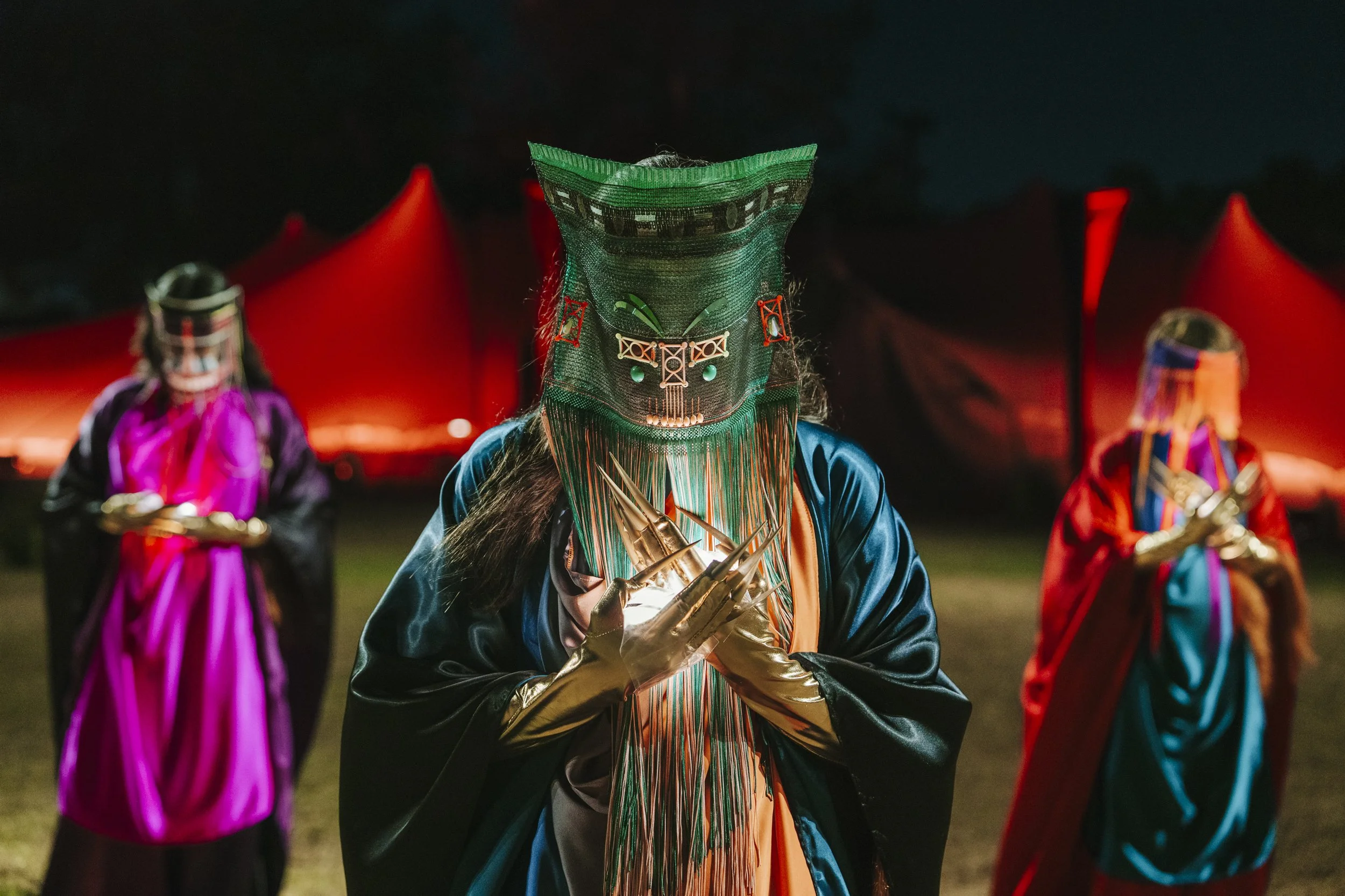 Three archetypal figures adorned in regalia including ceremonial masks, gold gloves, and ornate accessories, performing purposeful gestures at an outdoor cultural event at night.