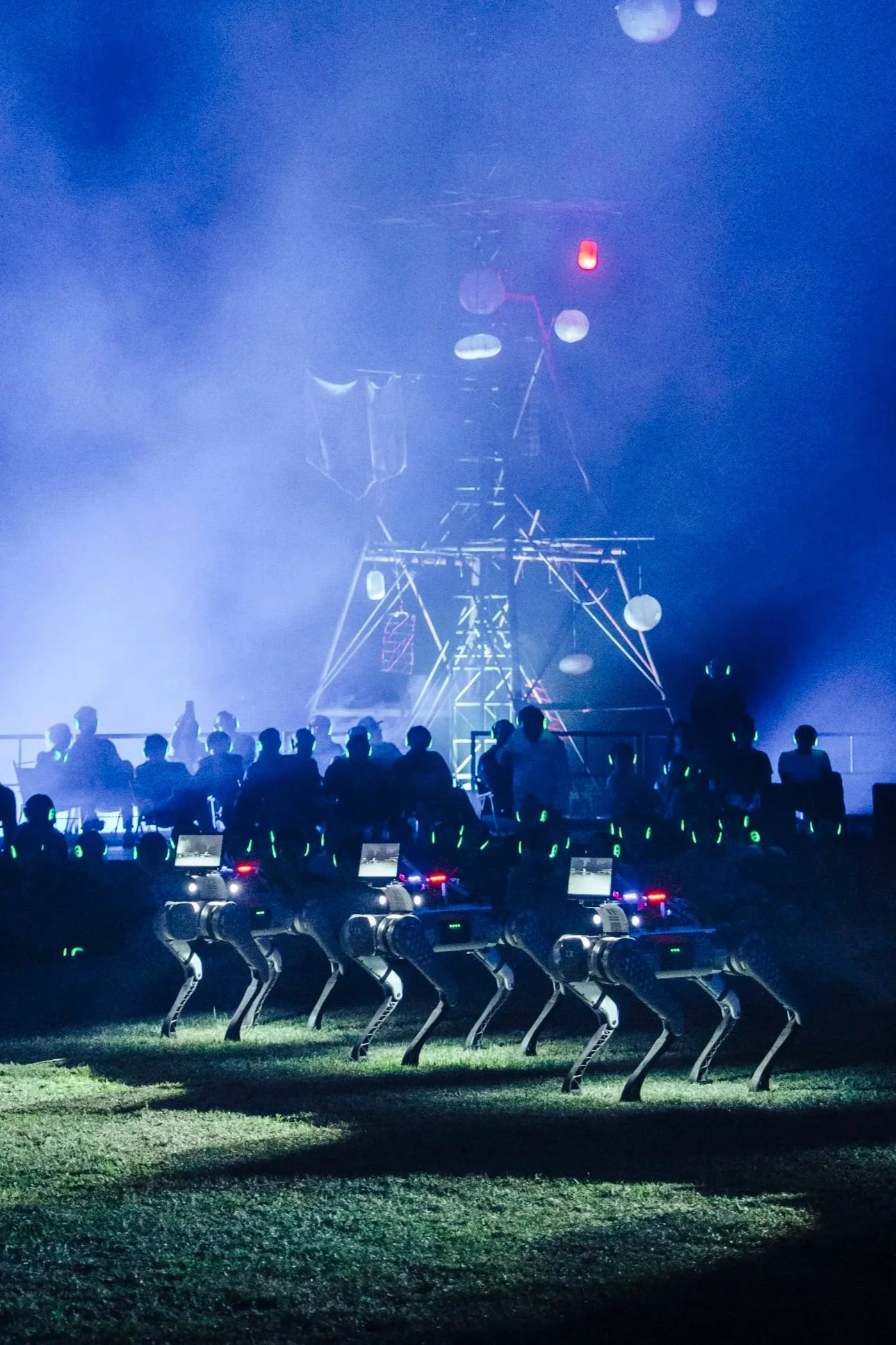 An outdoor stage with lighting and fog effects, with a group of audience members. In the lower part of the image, there are robotic quadruped figures standing menacingly in a line.