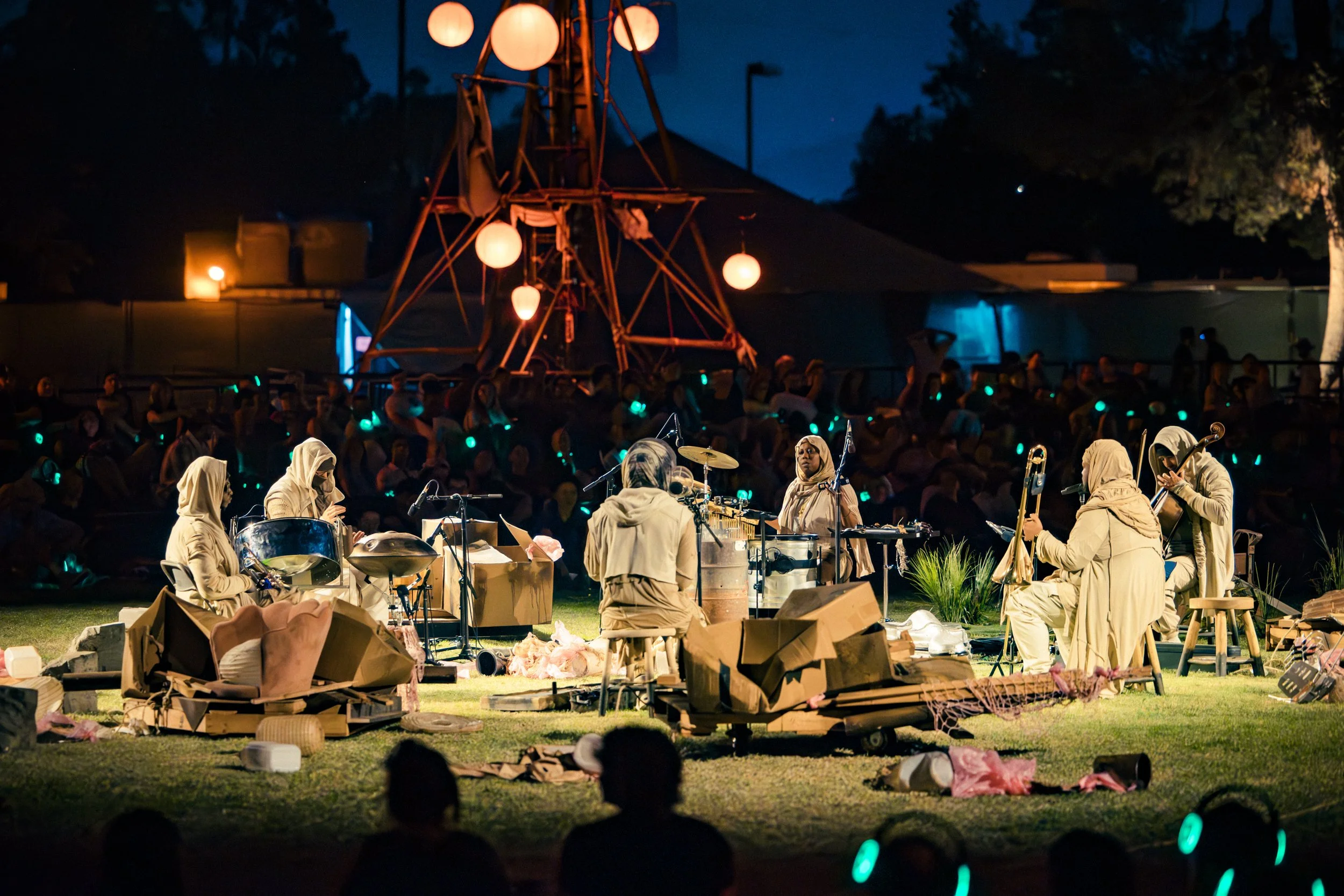 A nighttime outdoor performance by The Immeasurables, adorned in beige costumes and playing instruments among a smoldering pile of debris. The audience is seated in front of the stage. The background features trees and a dark sky.