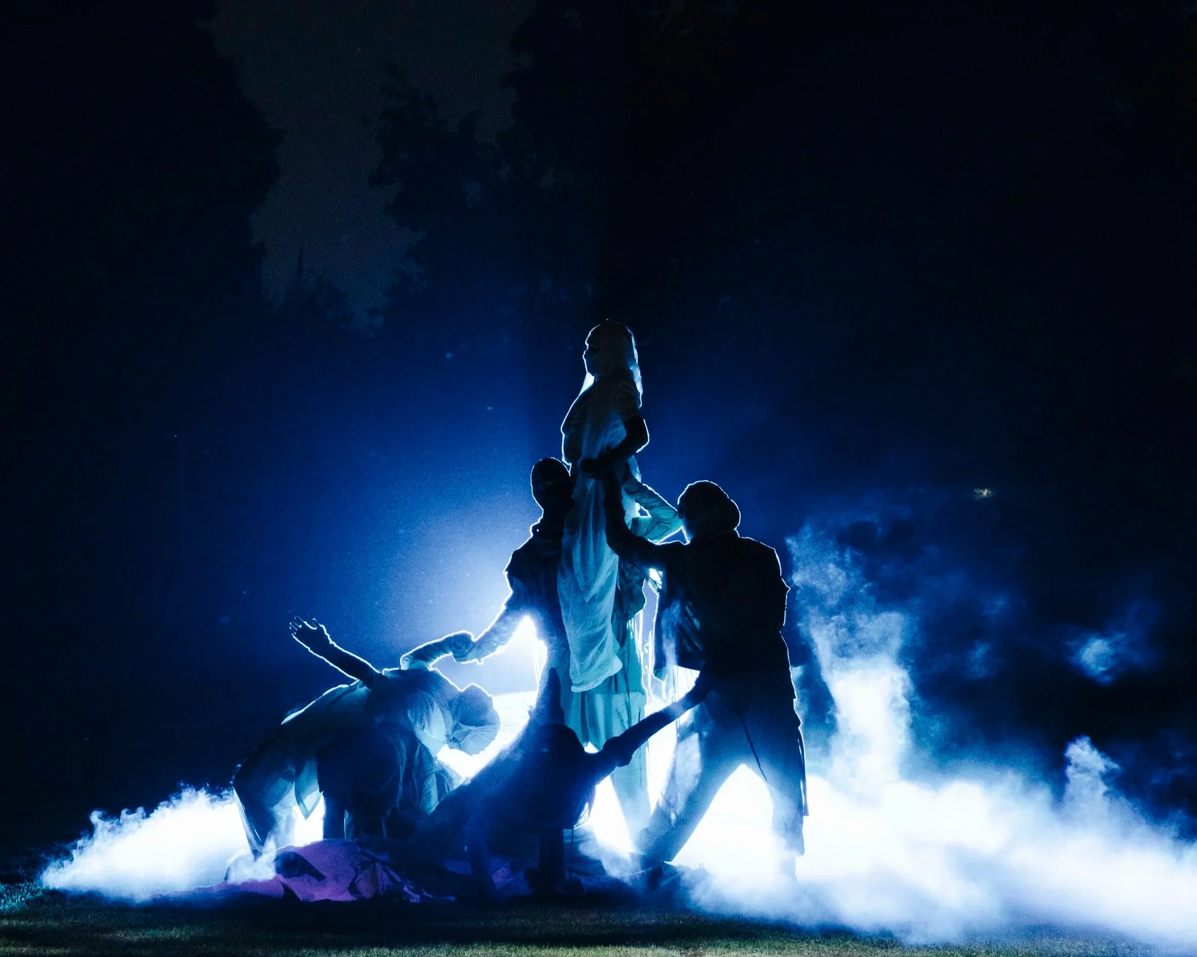 Silhouette of a group of dancers forming a human pyramid in front of a bright light and SFX under a night sky with clouds.