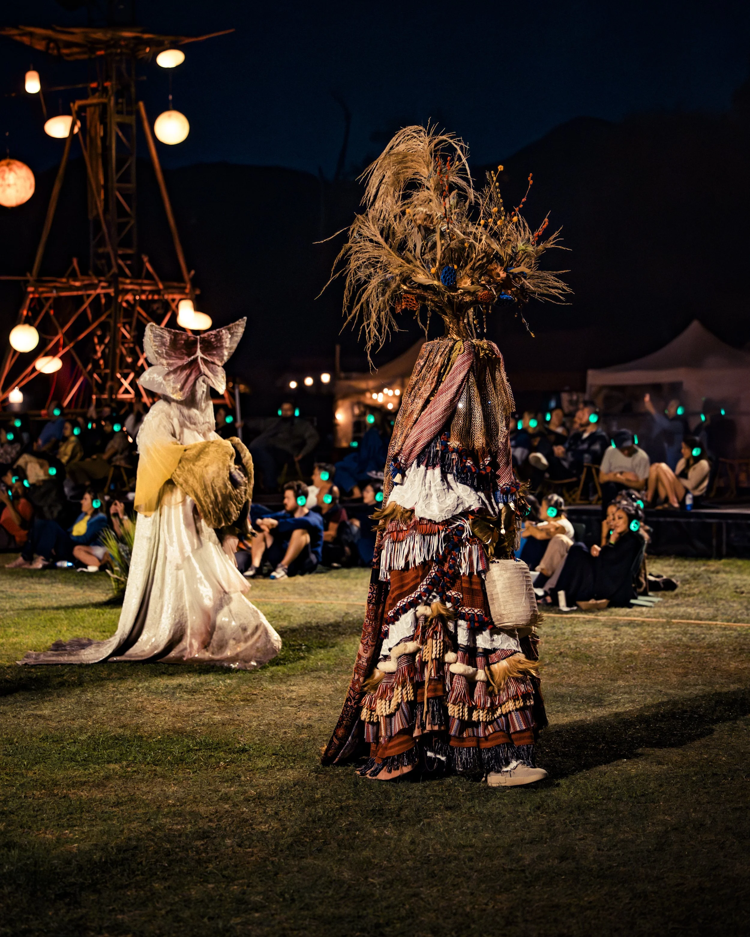 Two earthly figures in elaborate costumes and masks, decorated with feathers and fabric, walking on an outdoor stage. A large tower with hanging lanterns and a crowd with people wearing headphones is visible in the background.