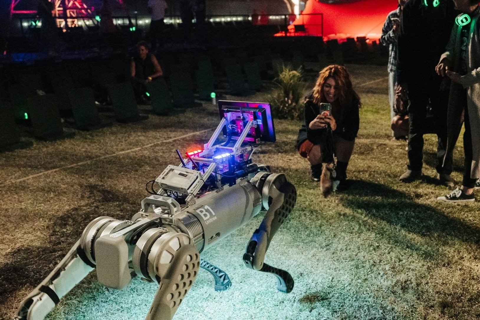A robotic dog with a metallic body and legs, equipped with electronic components, stands outdoors on grass at night, while a woman captures a selfie in the mirrored screen.