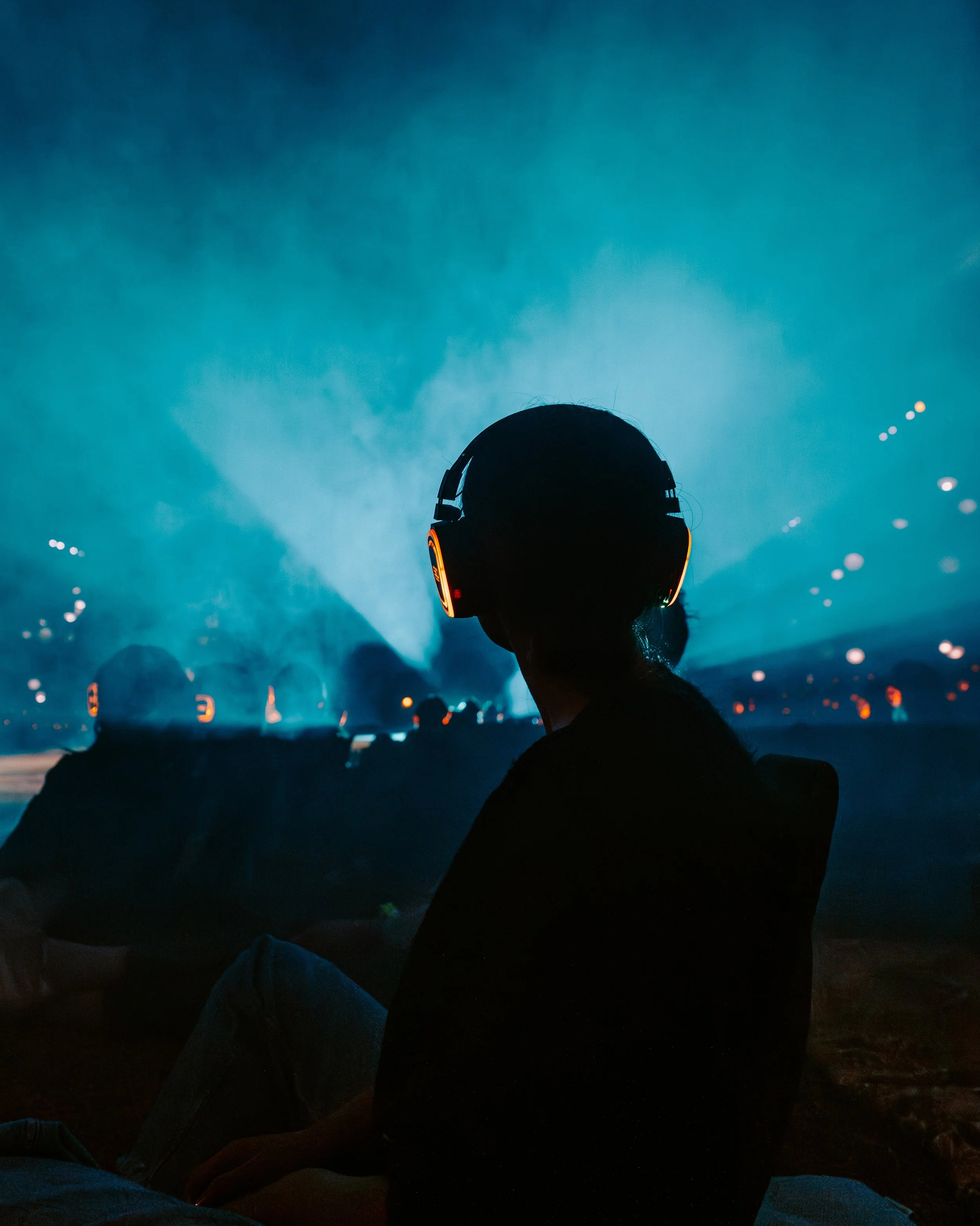 A person with silent disco headphones is sitting outdoors at night, watching a drone art display in the sky.