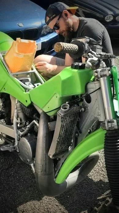 A man wearing sunglasses and a backward cap working on a green motorcycle with its body panels removed, showing parts like the radiator and frame, in a parking lot.