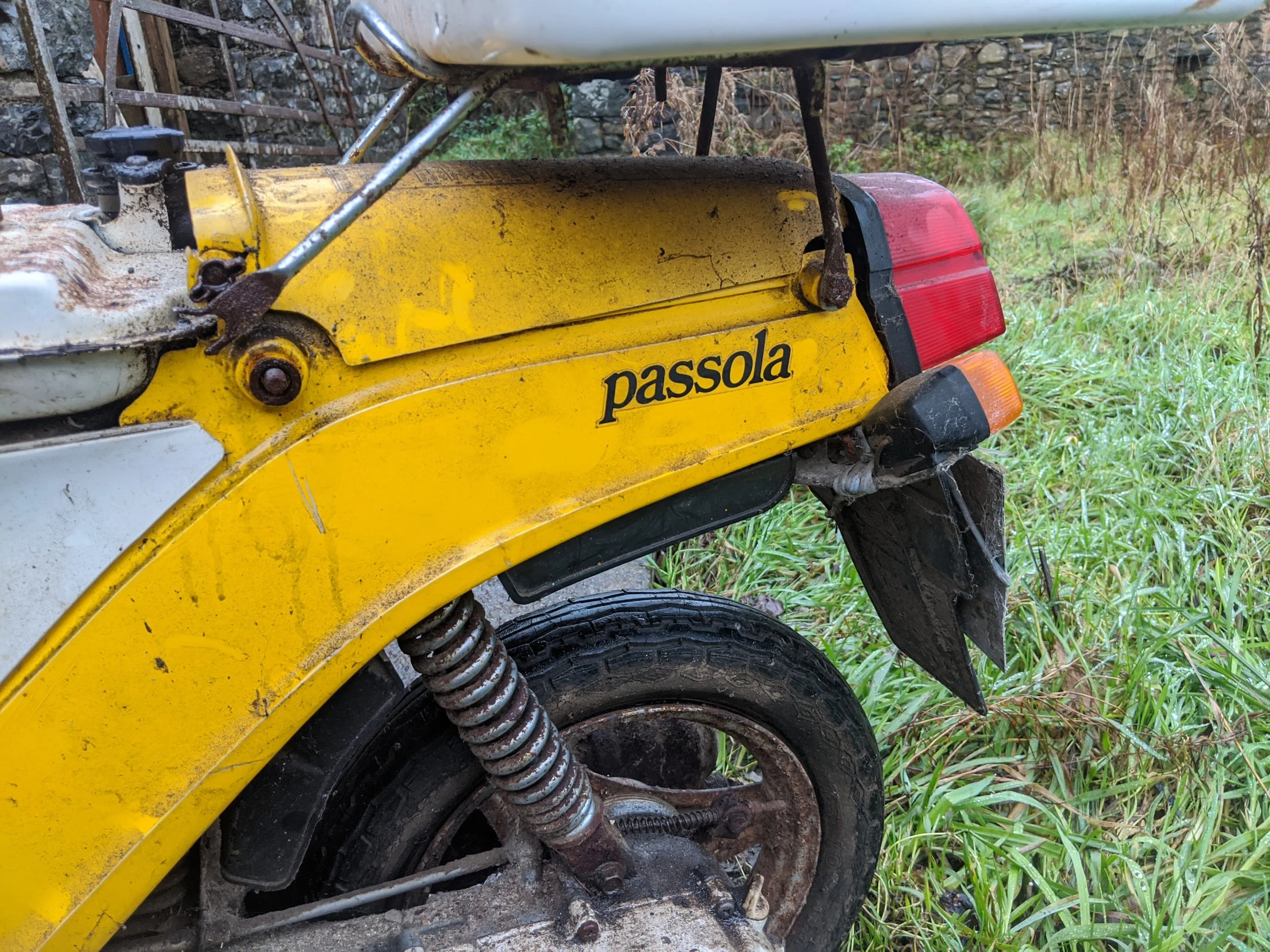 Close-up of an old, yellow Passola moped with rust on parts of the frame and suspension, parked on grass near a stone wall, with forest in the background.