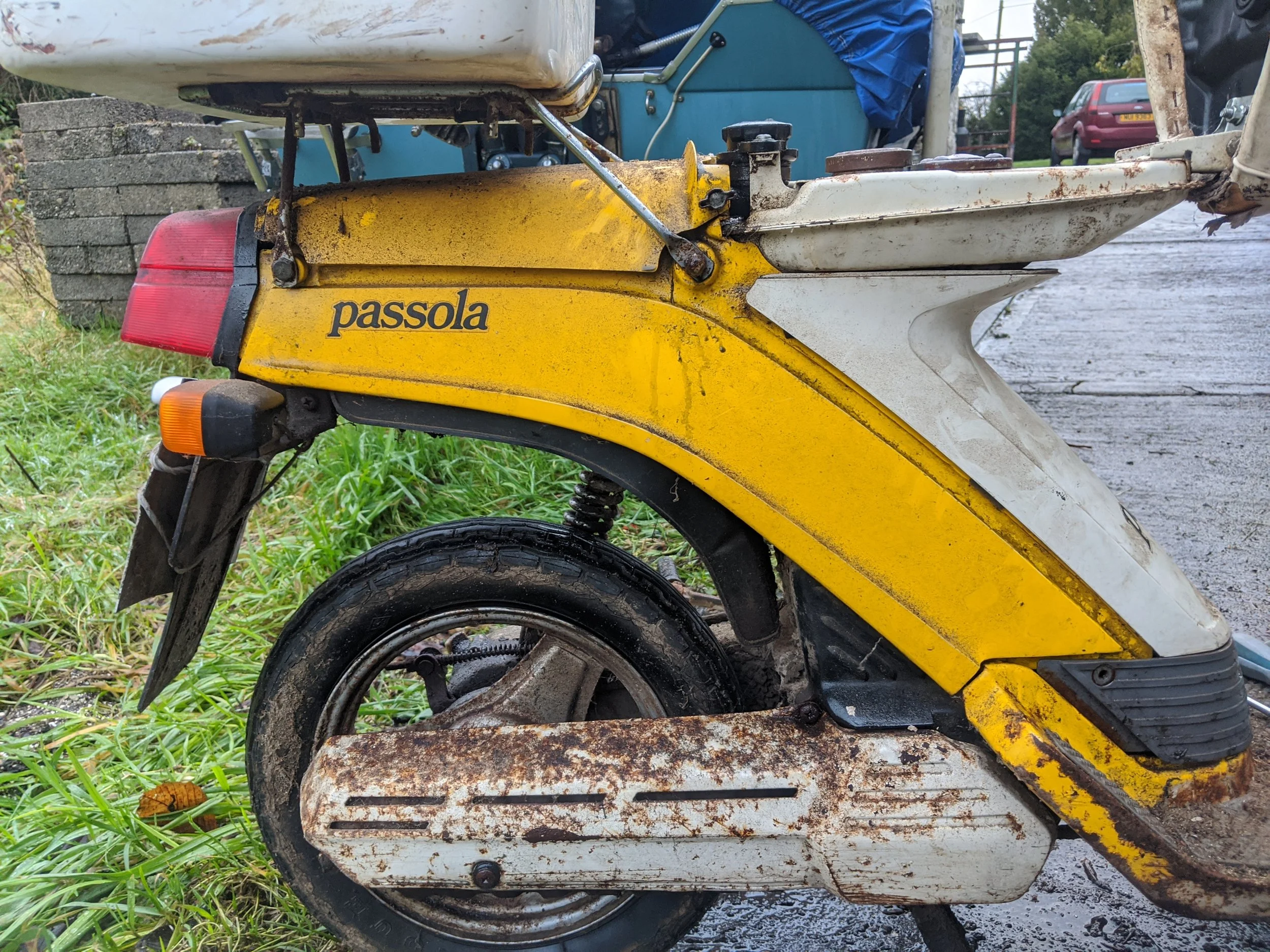 Close-up of a vintage yellow Passola motor scooter with rust and dirt, parked on grass and dirt, with a brick wall, blue tarp, parked cars, and trees in the background.