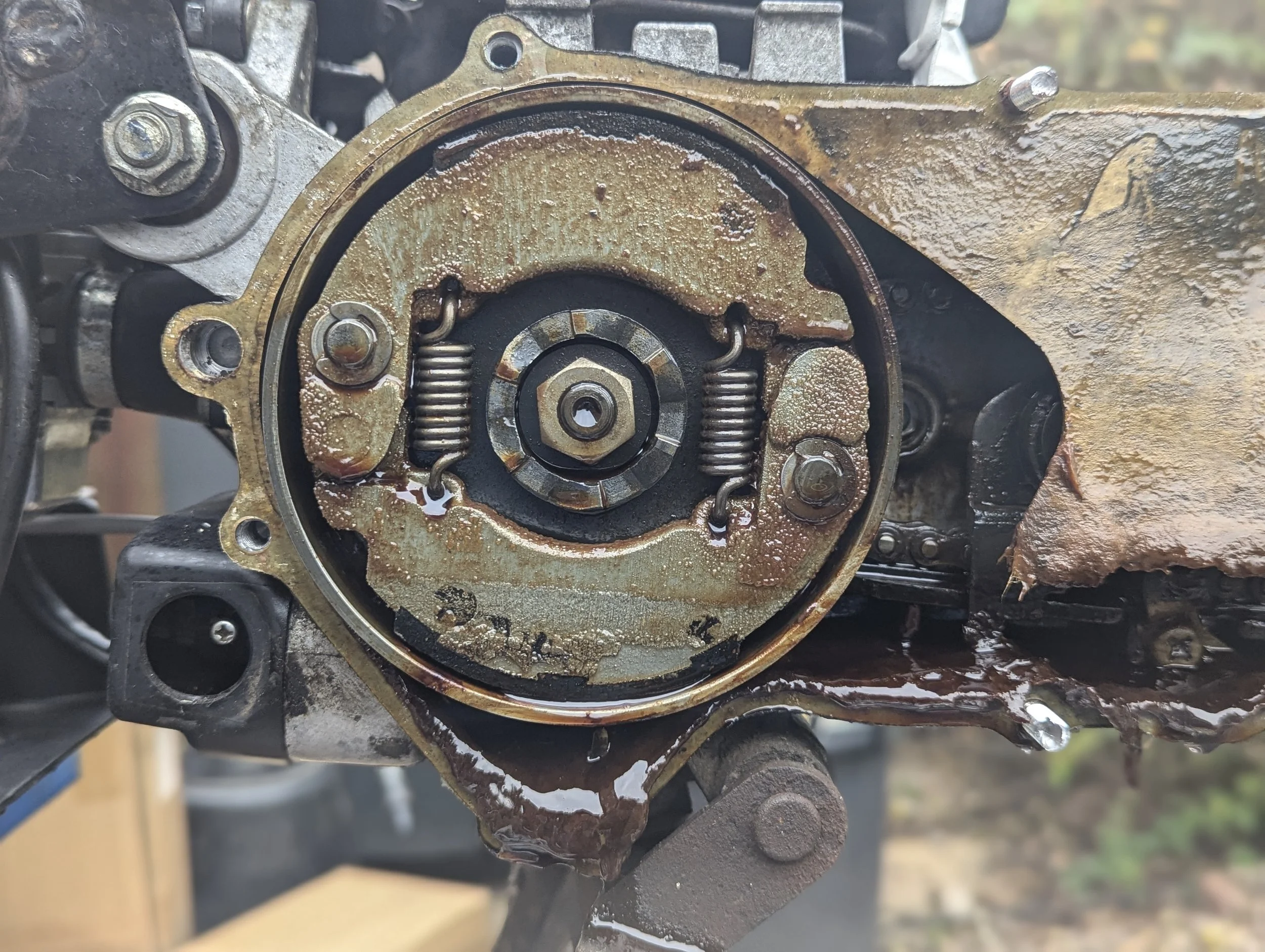 Close-up of an exposed motorcycle engine part with oil and grime, showing springs, bolts, and metal components.