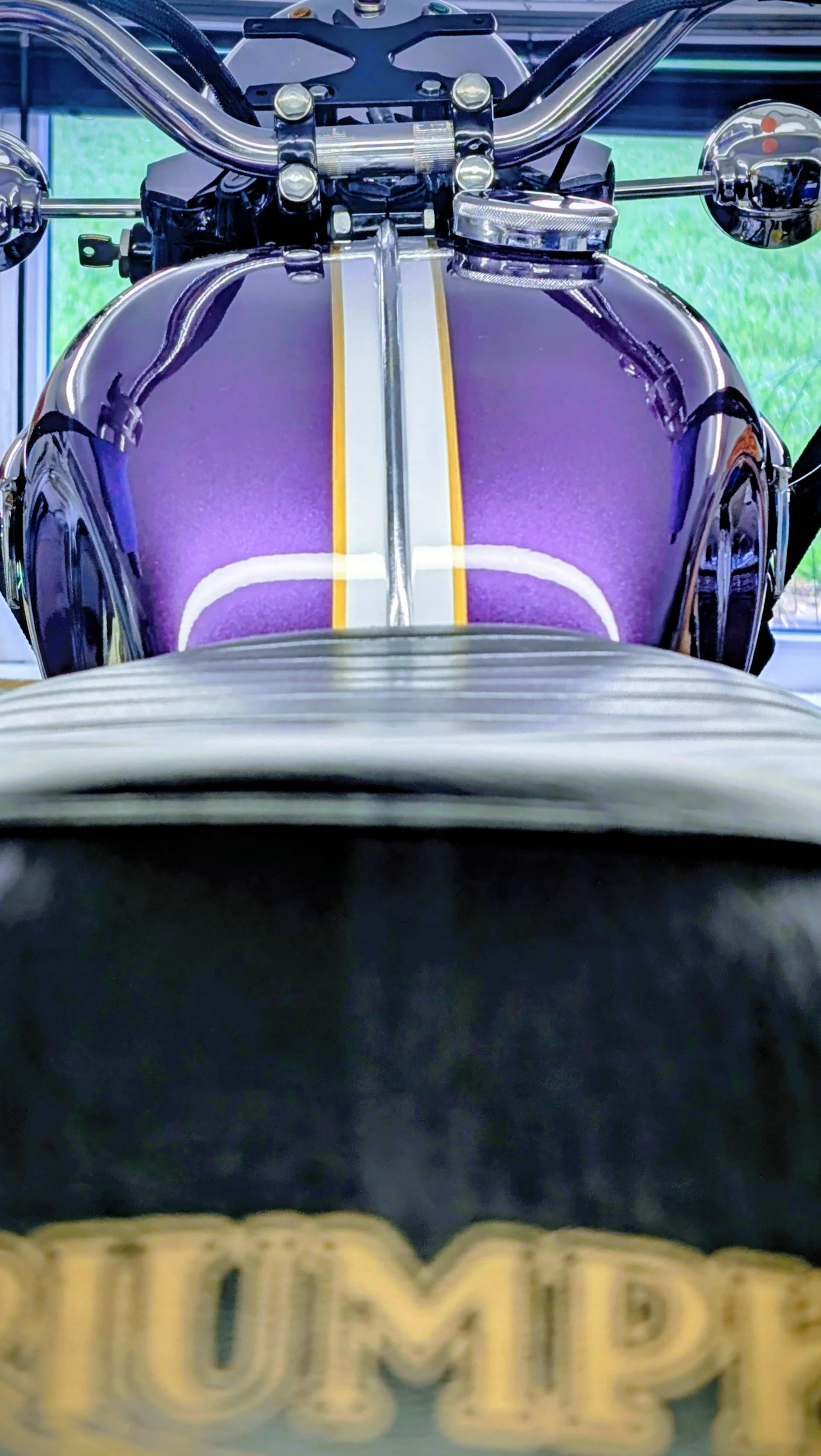 Close-up photo of a vintage motorcycle's front end, showing the shiny chrome handlebars, purple fuel tank with yellow and white stripes, and a blurred yellow logo at the bottom. Original Triumph vintage colours, restored. Barn find Triumph T100 resto