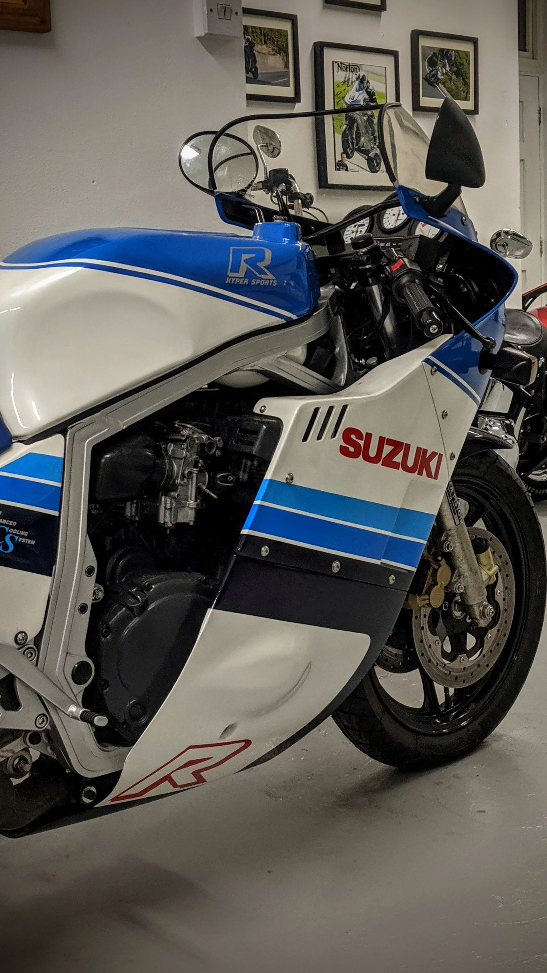 A vintage Suzuki motorcycle with a custom blue, white, and red racing livery parked indoors, with framed racing photos on the wall behind it.