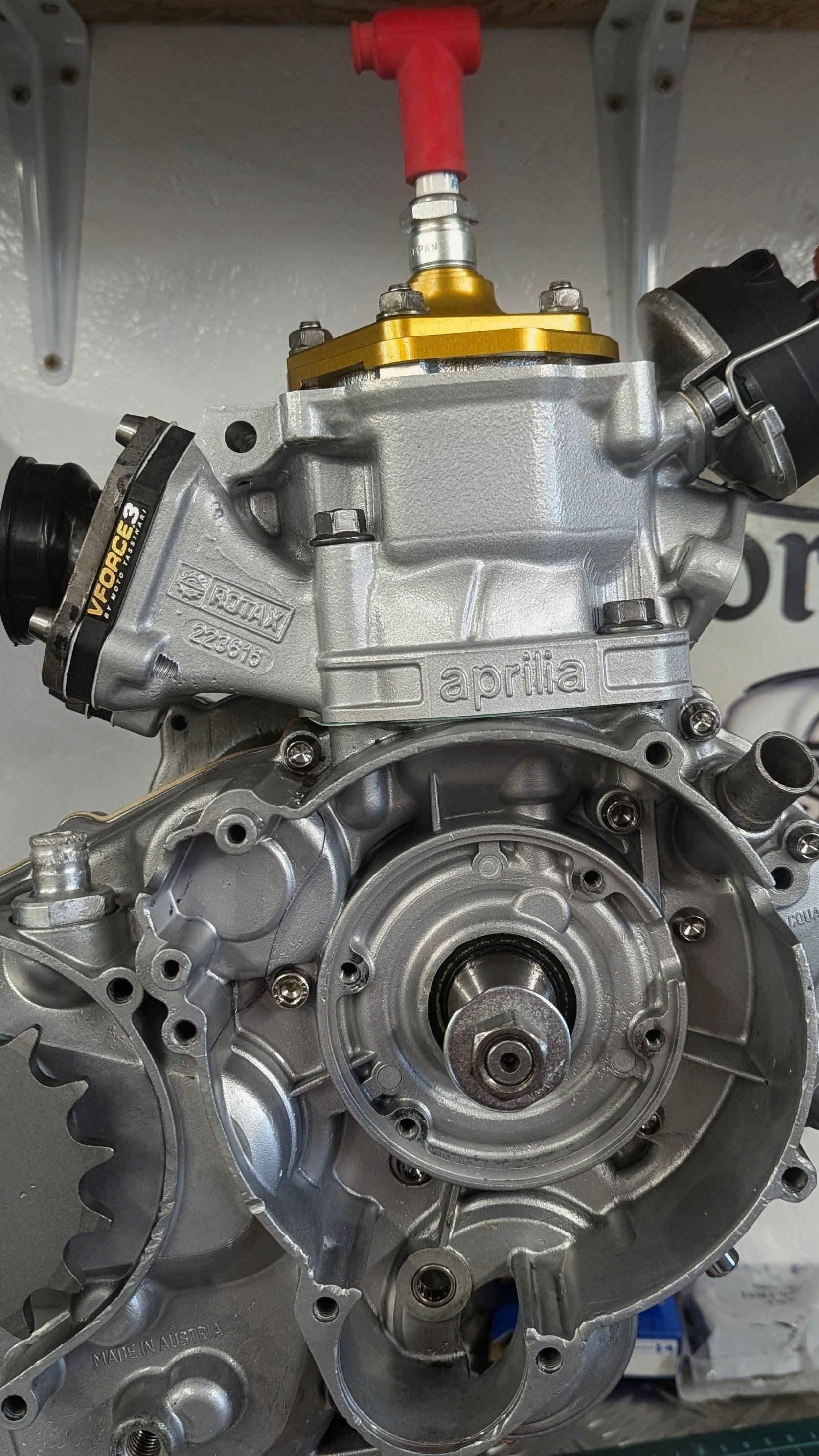 Close-up of a silver motorcycle engine with gold accents and black components, branded 'aprilia', mounted on a workbench.
