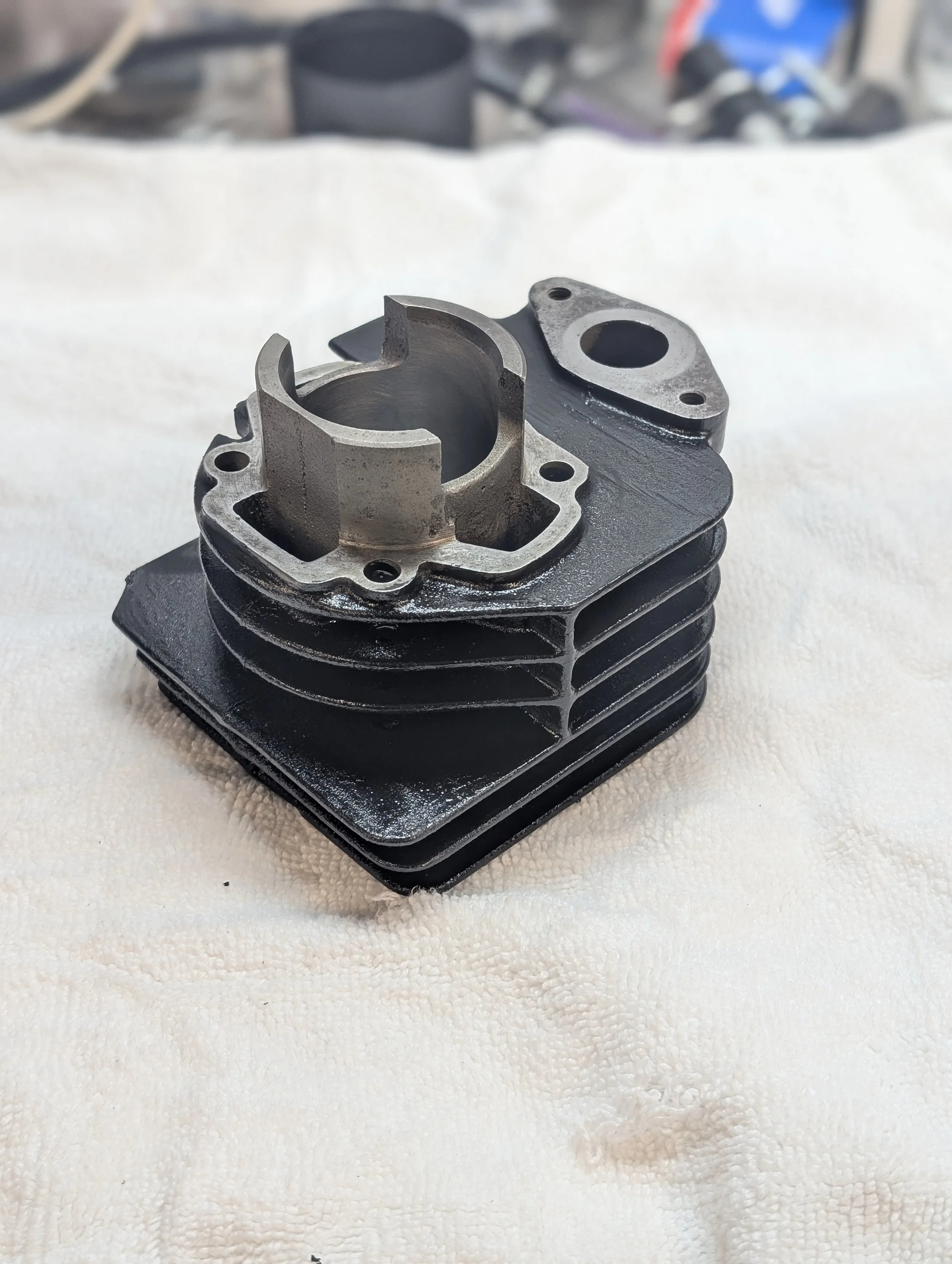 A metal engine cylinder with cooling fins and bolt holes resting on a white textured surface, with a blurred background of tools and parts.