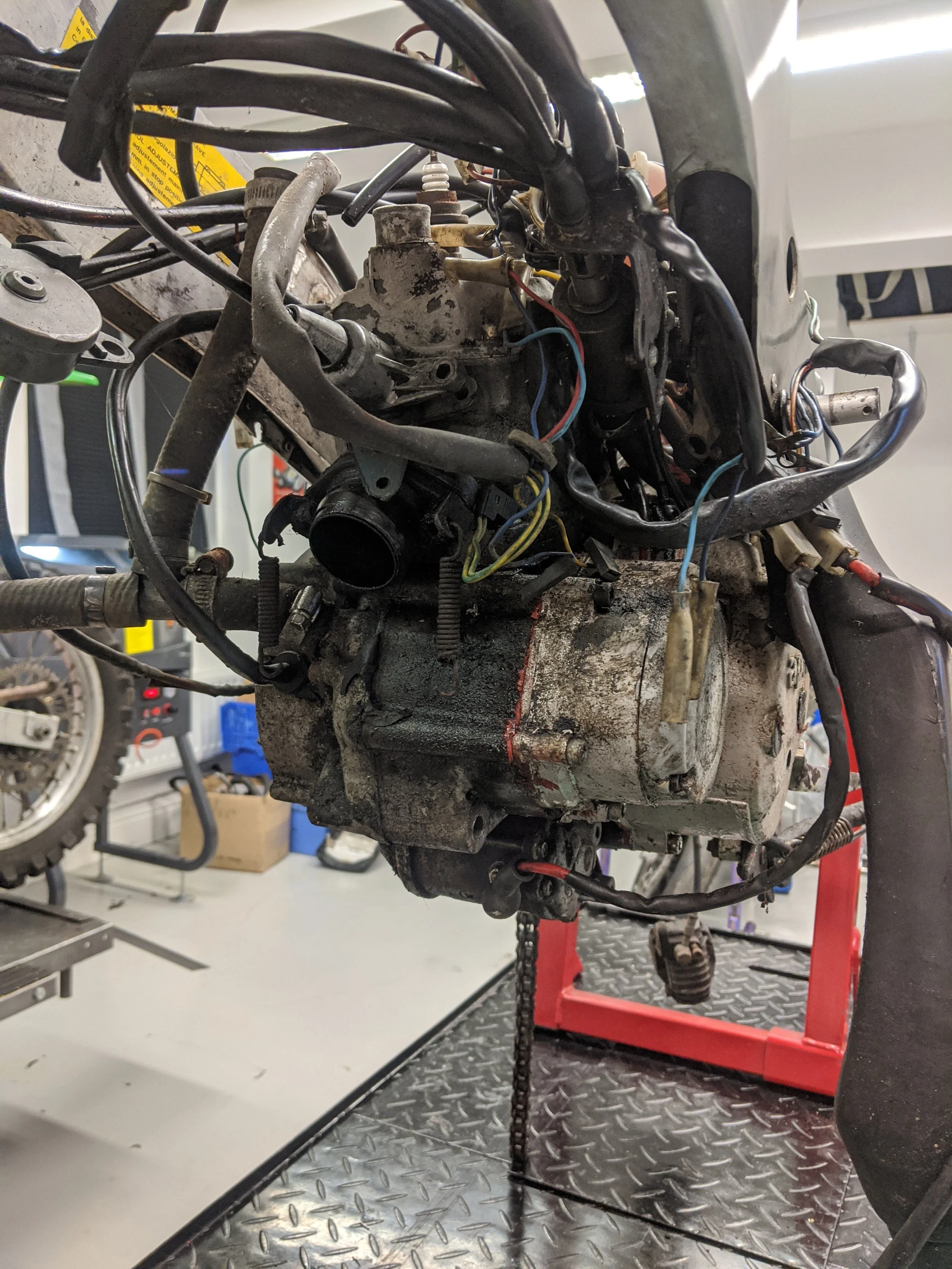 Close-up of a motorcycle engine and frame with wires and components, mounted on a red stand inside a garage or workshop with tools and equipment in the background.