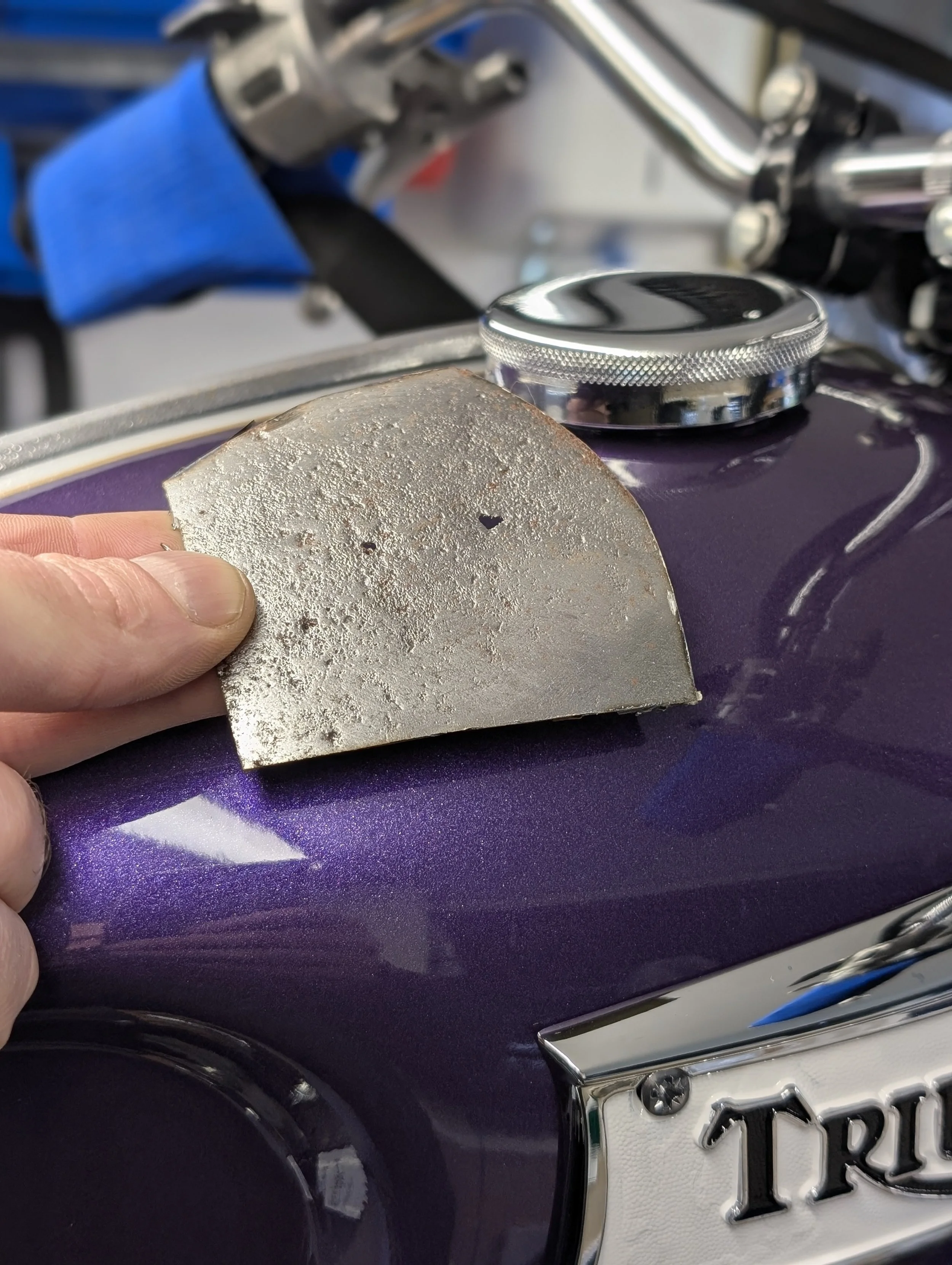Close-up of a motorcycle fuel tank with a shiny chrome fuel cap, a purple paint job, and a black and silver license tank badges with partial text visible showing Triumph. The background shows a person's hand holding a small, rough piece of metal whic